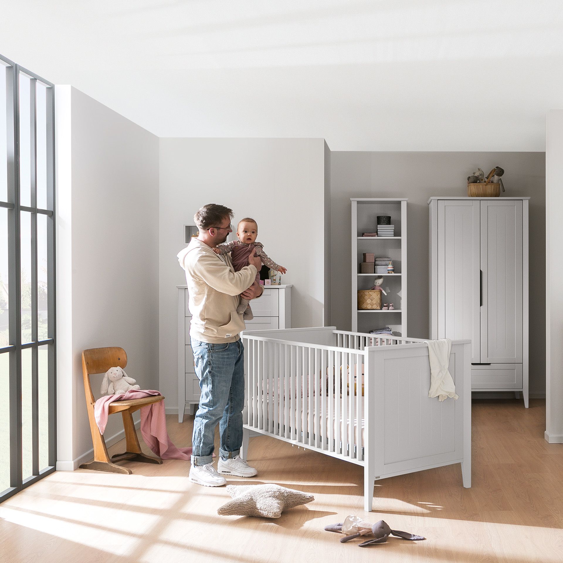 A man holds a baby beside a crib in a bright, modern nursery room.