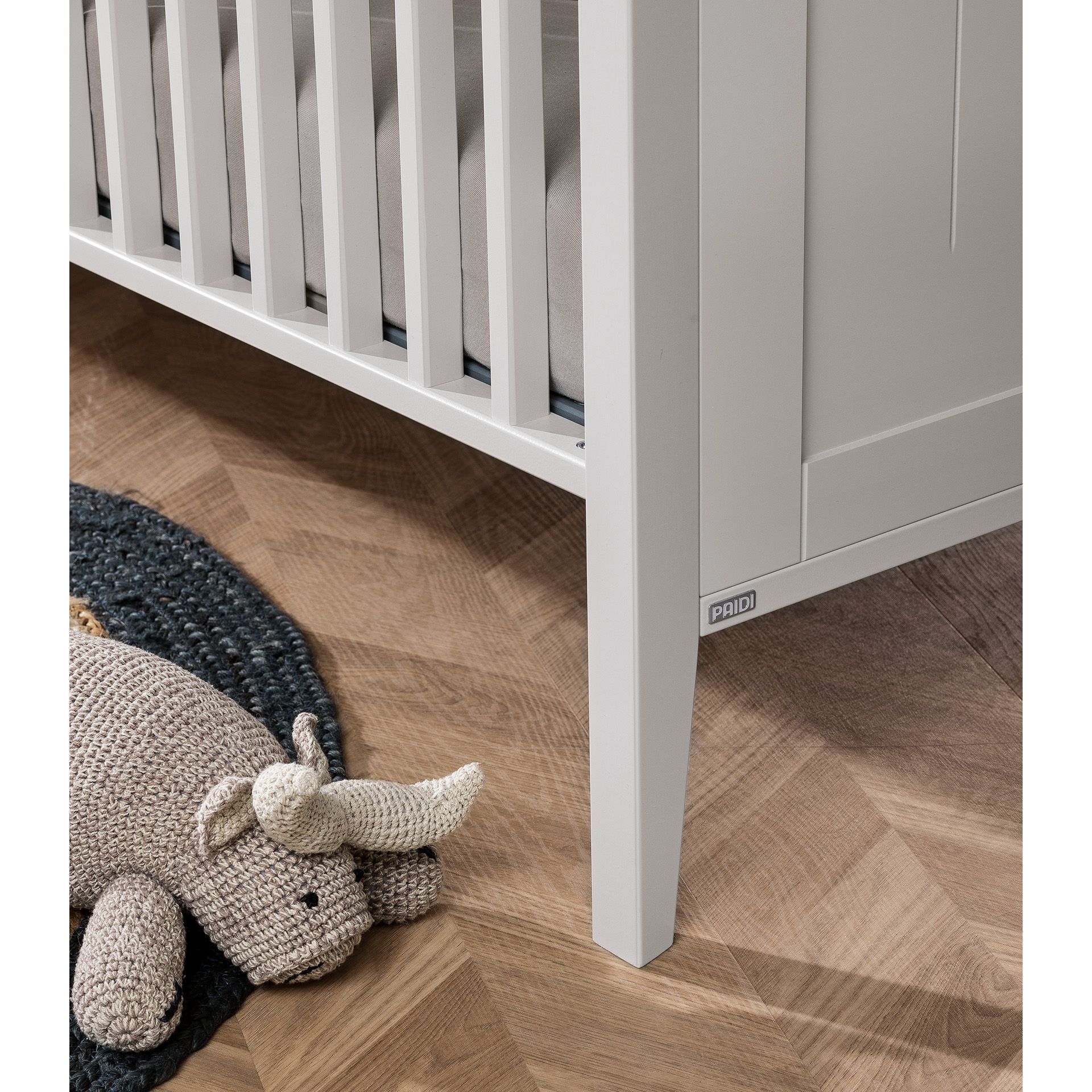 A knitted rhino toy lies beside a white crib on a wooden floor.