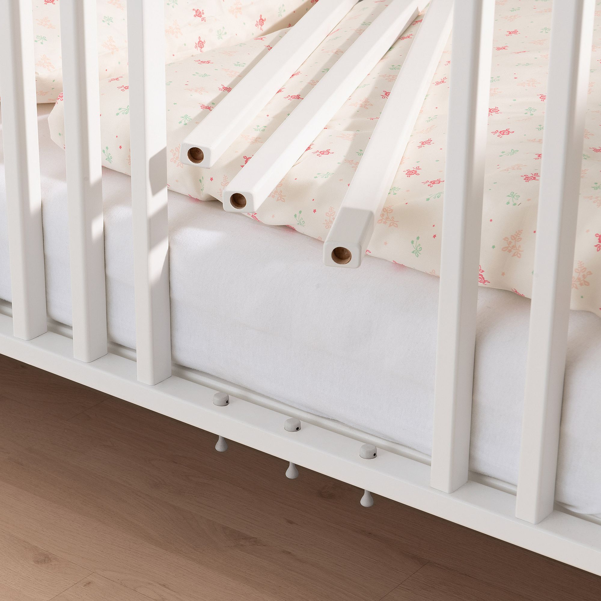 Weißes Kinderbett mit geblümter Bettwäsche, eine Seite ist abgenommen, sodass der Holzboden sichtbar ist.