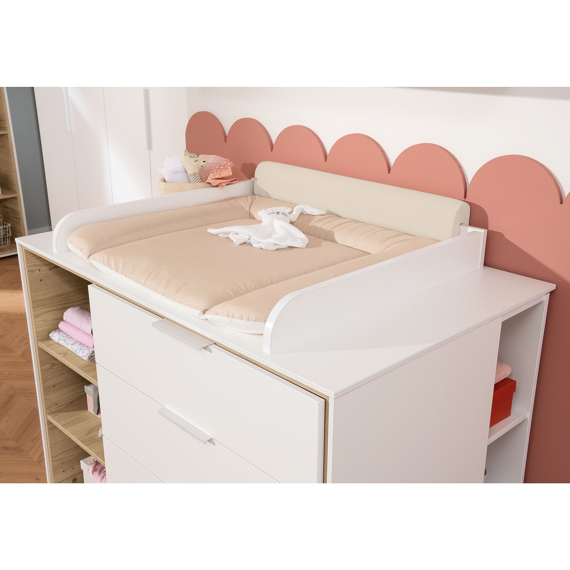 Weißer Wickeltisch mit beiger Unterlage, Babykleidung und rosafarbenem Wanddekor mit Wellenschliff.
