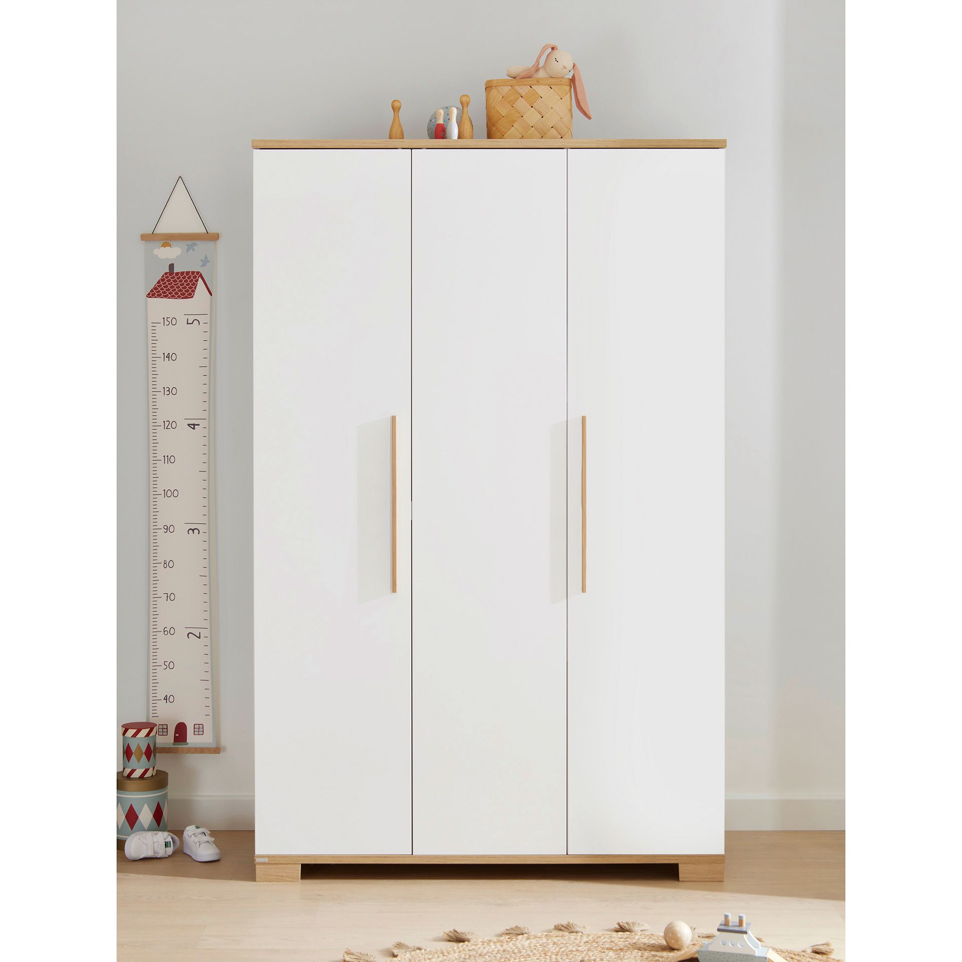 Drehtürenschrank Yolanda mit 3 Türen Weißer Kleiderschrank mit Holzgriffen, Höhentabelle und Spielzeug auf hellem Holzboden.