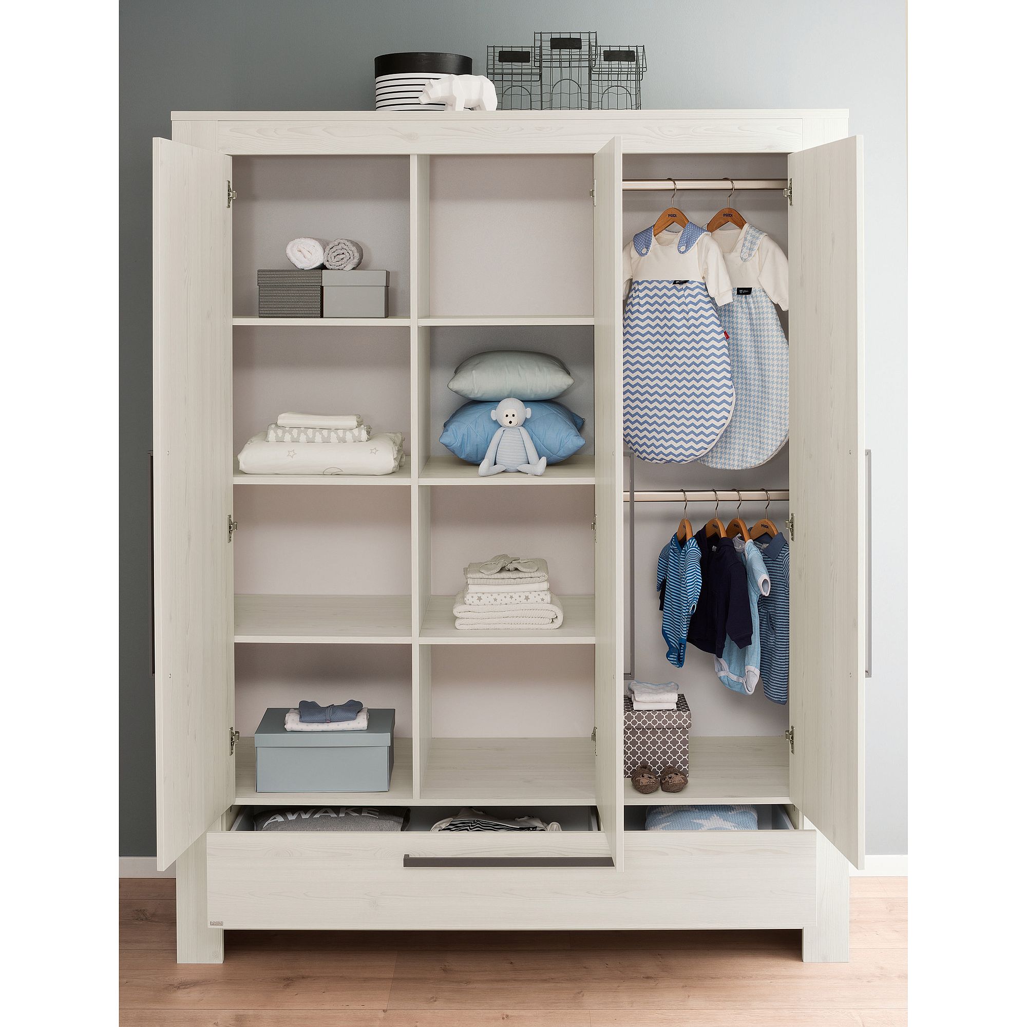 Drehtürenschrank Laslo mit 3 Türen und 1 Schublade Weißer Kleiderschrank mit offenen Türen, darin ordentlich geordnete Babykleidung und Spielzeug.