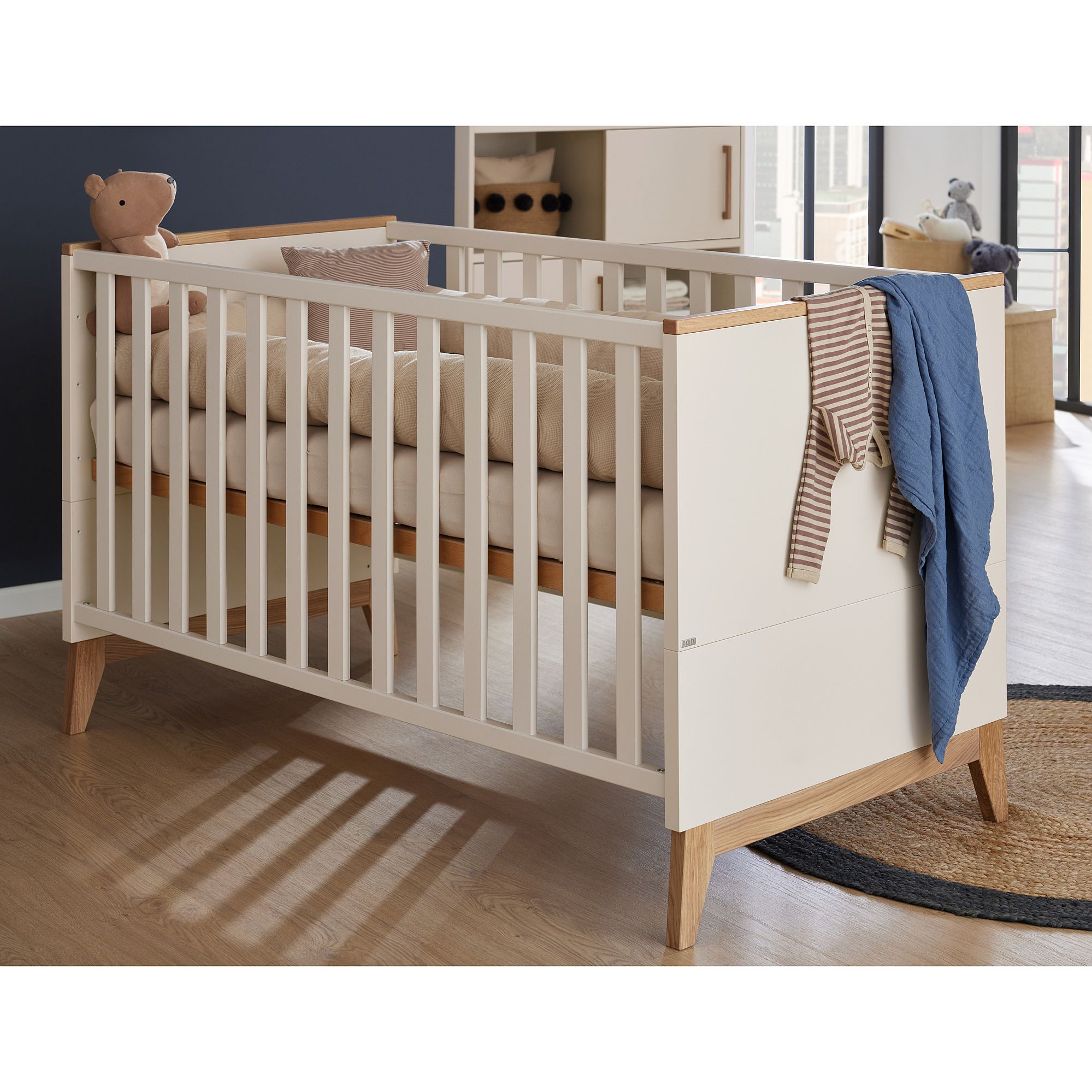 Ein modernes Babybett mit Bettzeug, einem Teddybär und an der Seite hängender Kleidung.