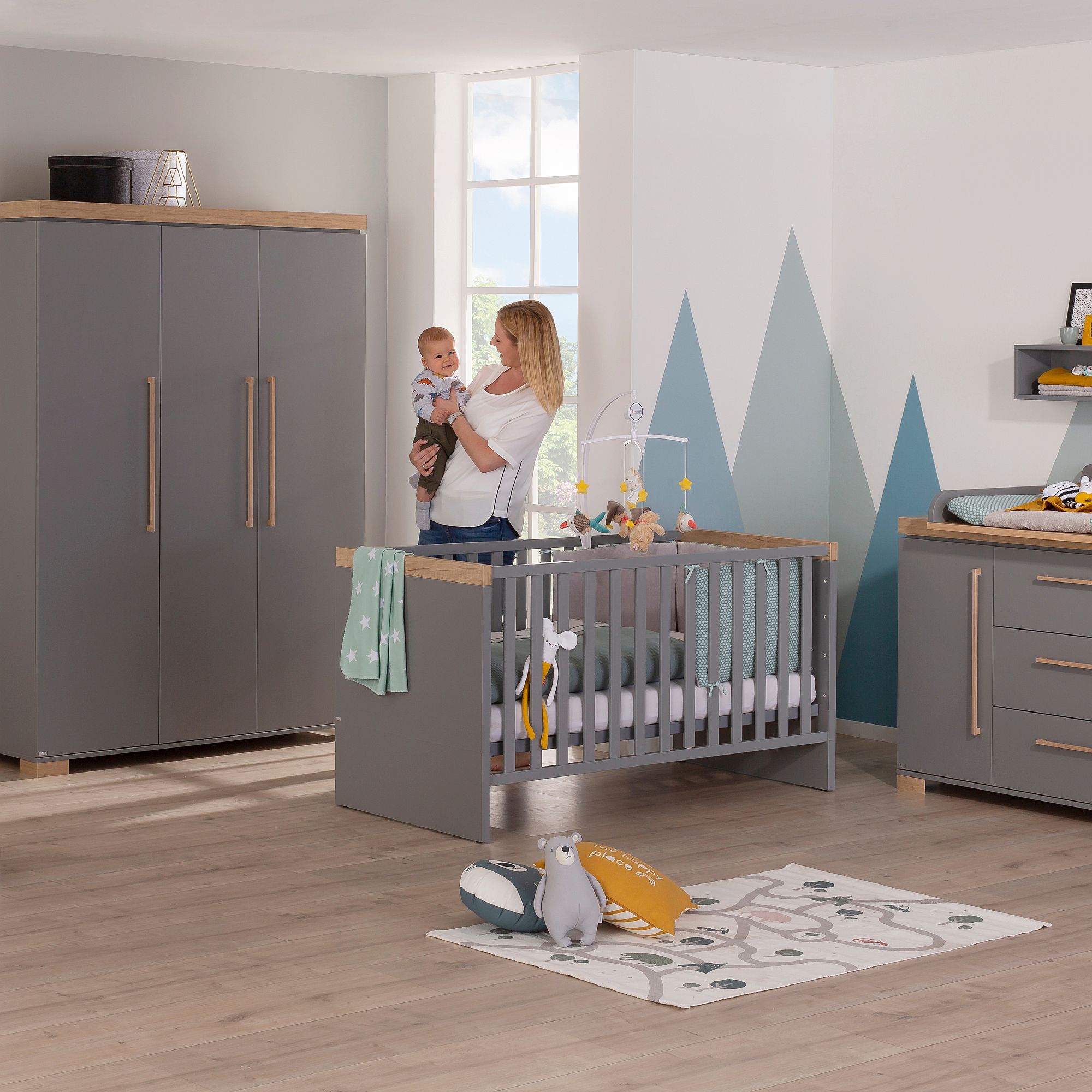 Eine Frau hält ein Baby in einem modernen, in Grautönen gehaltenen Kinderzimmer mit einem Kinderbett und Deko.