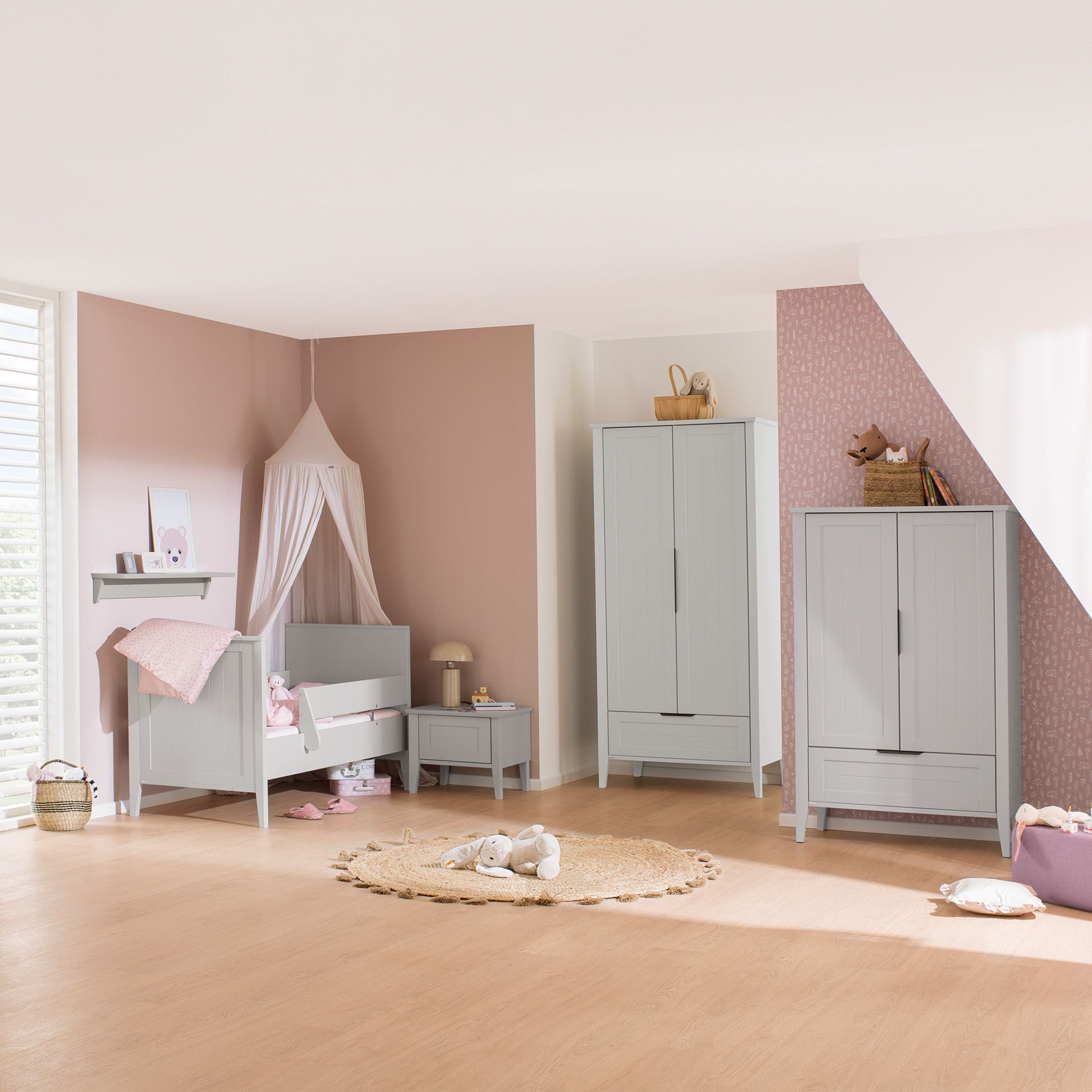 Nursery room with pastel pink walls, gray furniture, and toys on a round rug.