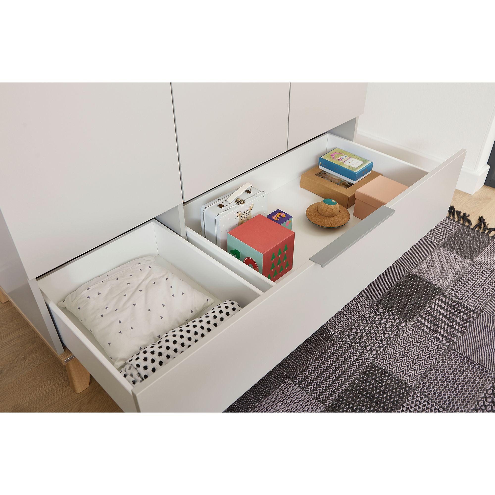 Drehtürenschrank Fiene mit 3 Türen und 1 Schublade Offene Schublade mit Büchern, Spielzeug, einem Kissen und einem gemusterten Teppich in der Nähe.