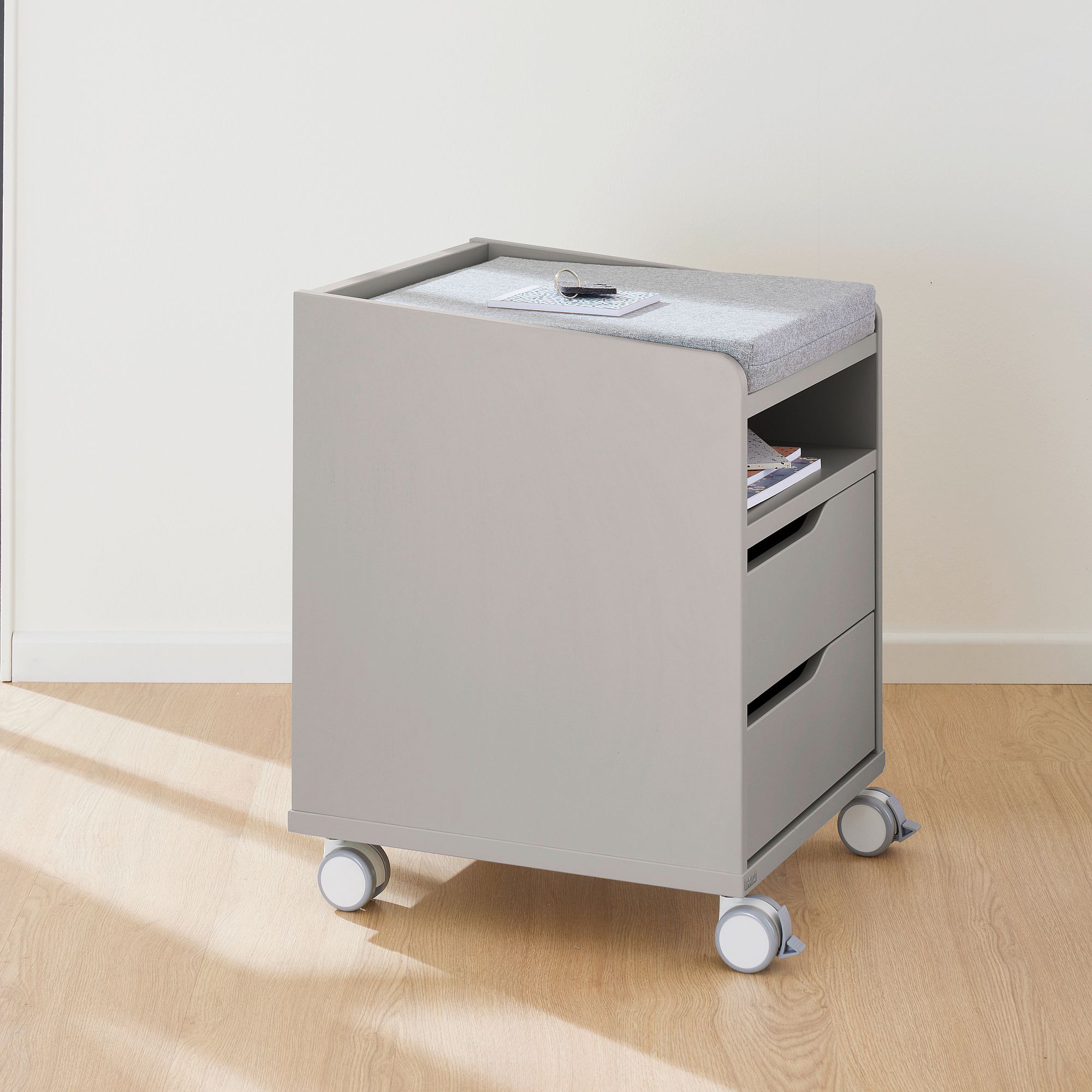 Ein grauer Rollschrank mit zwei Schubladen und einer gepolsterten Platte auf einem Holzboden.