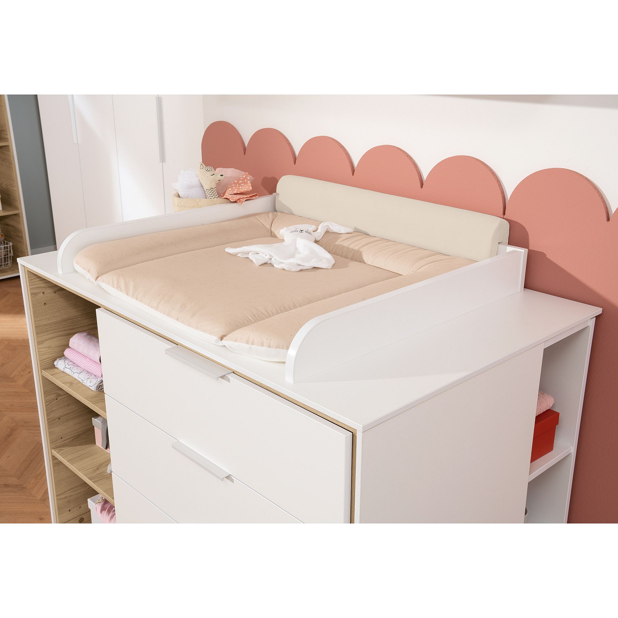 Weißer Wickeltisch mit beiger Unterlage, Kuscheltier und gefalteter Babykleidung darauf.