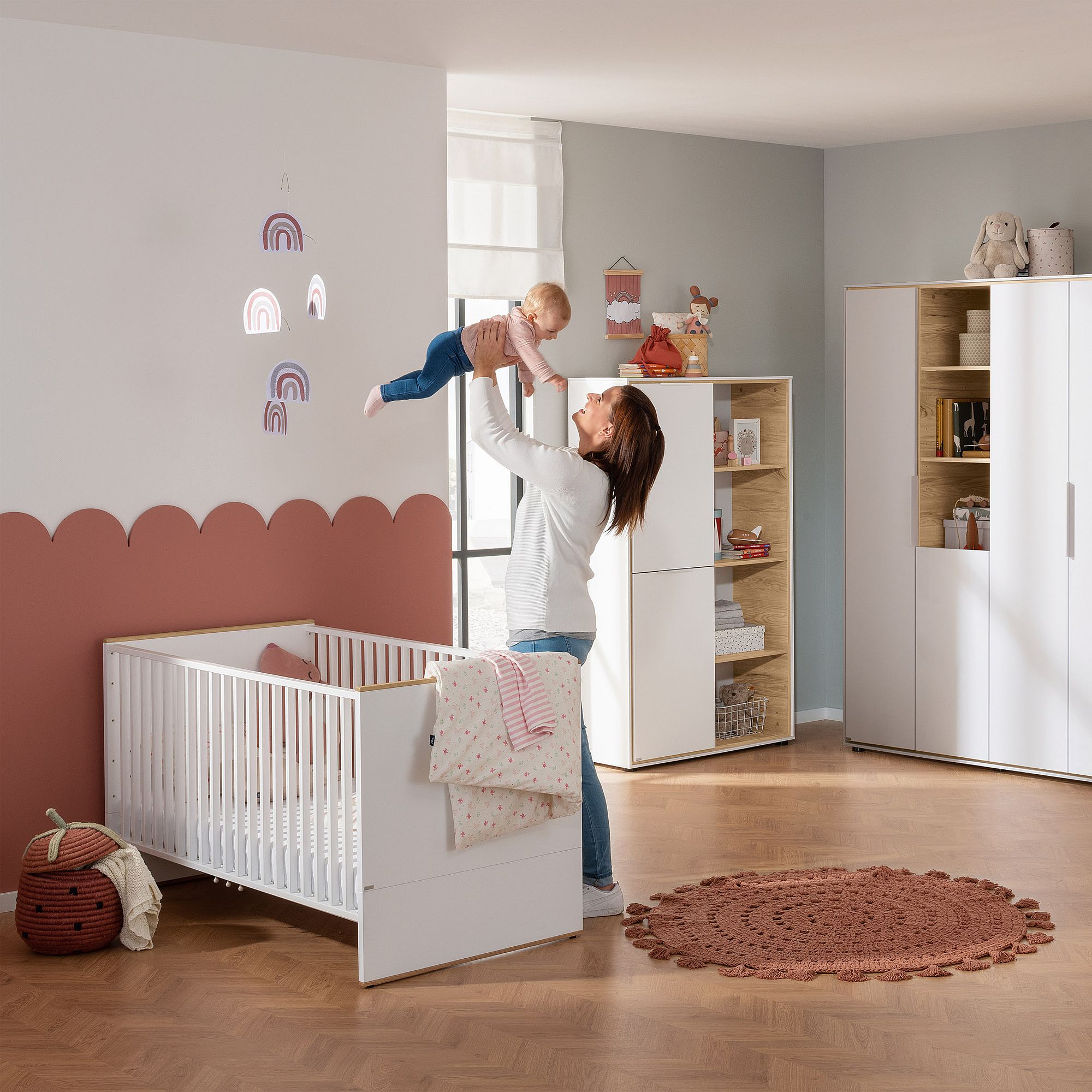 Eine Frau hebt ein Baby hoch in einem Kinderzimmer mit einem Kinderbett, einem Teppich und Regenbogen-Wandtattoos.
