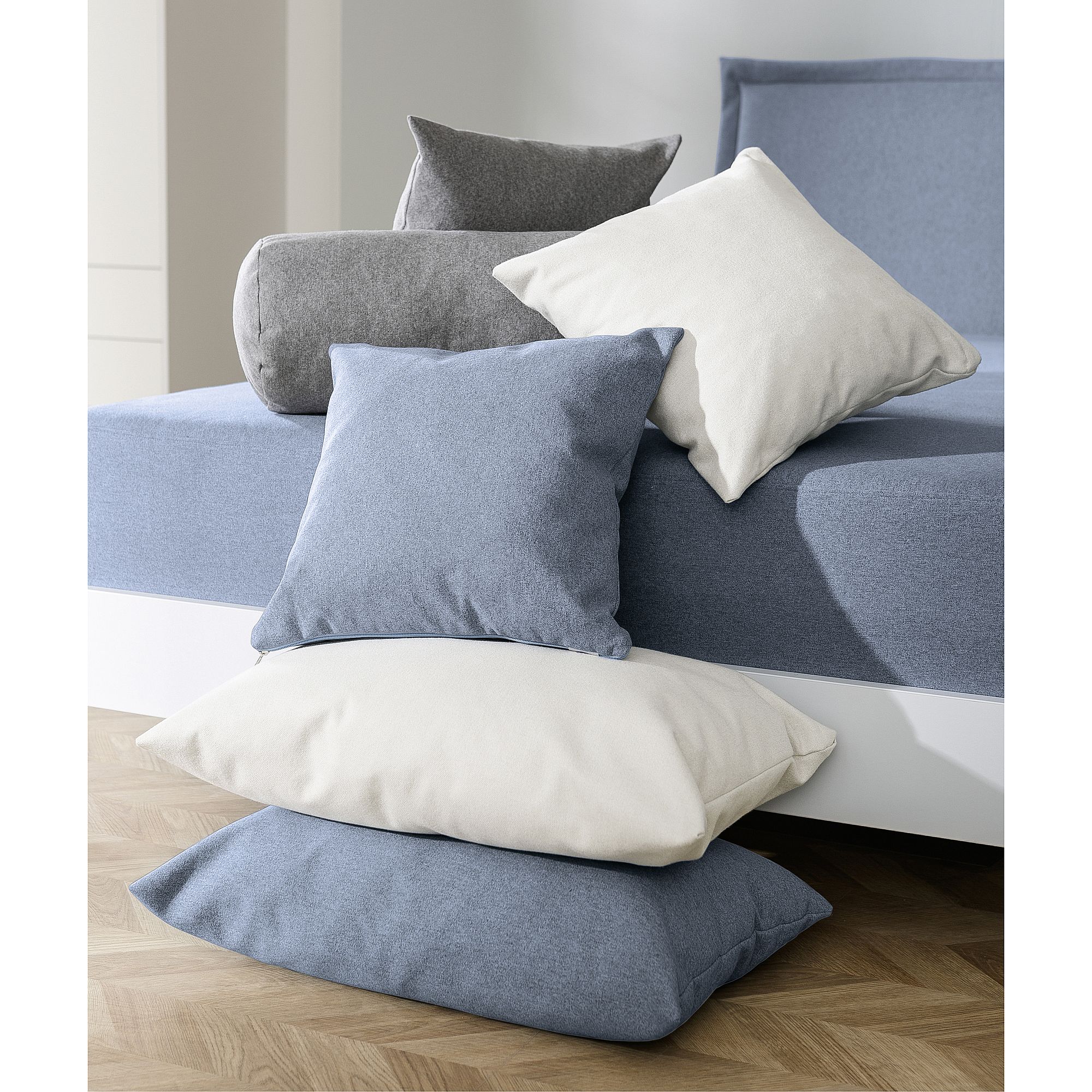 A stack of blue, gray, and white pillows next to a bed with blue bedding.