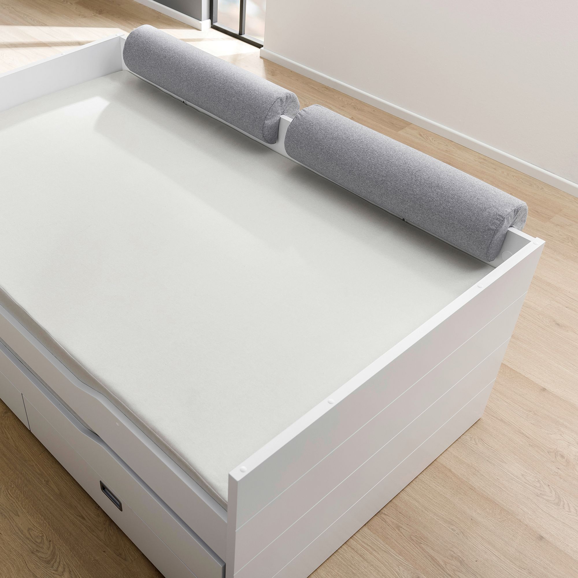 A white bed with a light gray cylindrical cushion along the headboard.