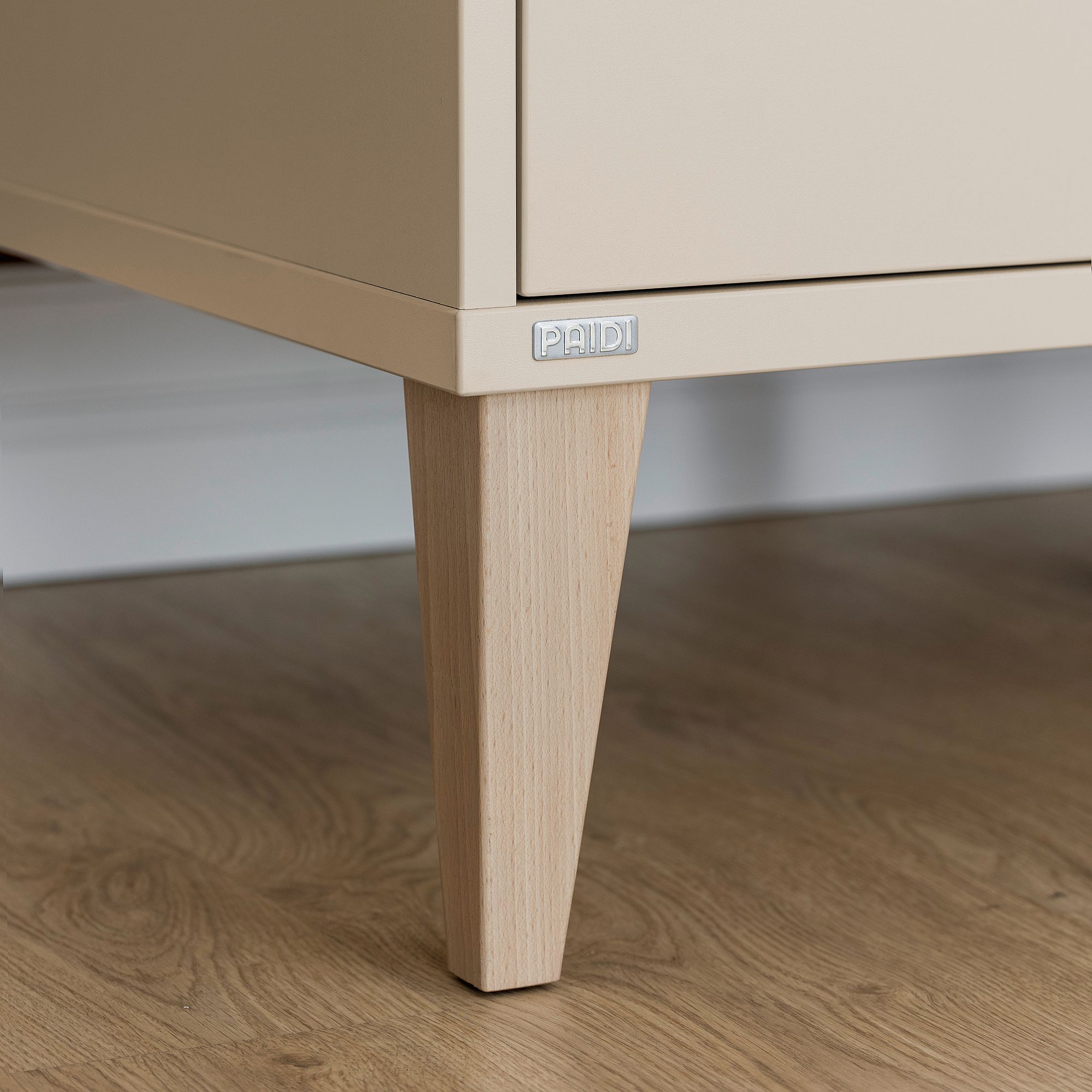 Beigefarbener Schrank mit Holzbeinen auf einem Hartholzboden, PAIDI-Etikett an der Ecke.