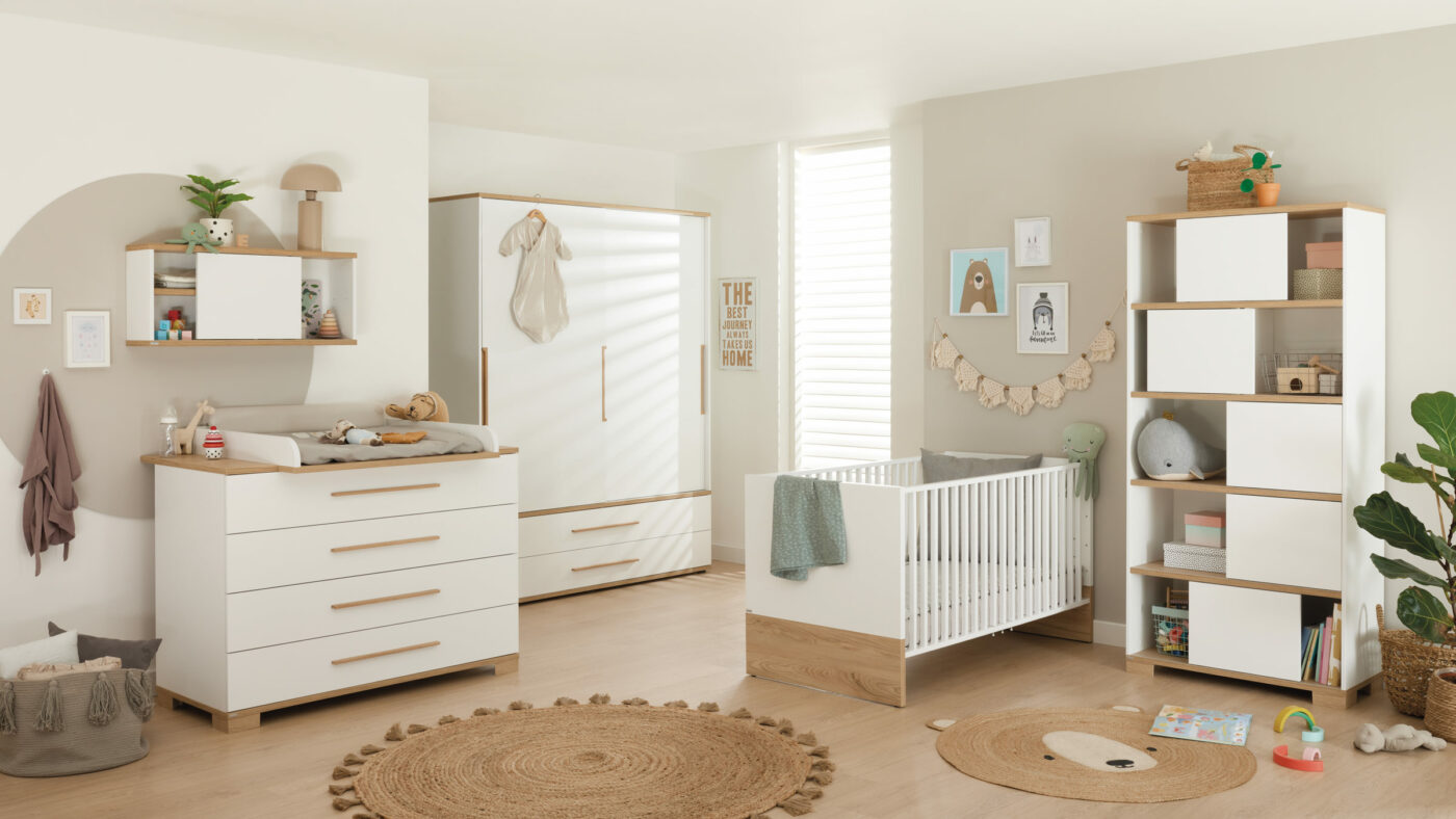 1101_Babyzimmer_paidi_yolanda_baby_0001-1400x788-ID1691208_37316279-7774-51f3-3d17-1c95e98d3d93.jpg Modernes Kinderzimmer mit weißen und hölzernen Möbeln, einem Kinderbett, einer Kommode, Regalen, Spielzeug und neutralem Dekor in einem hellen, gemütlichen Raum.