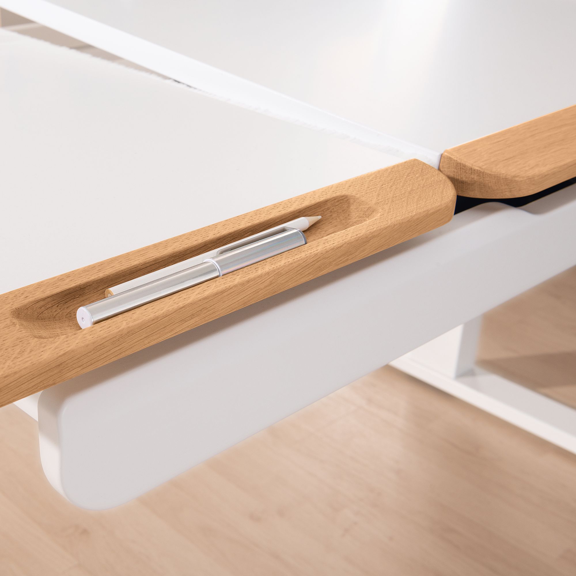 Ein weißer Schreibtisch mit einer Rille aus Holz, in der ein silberner Kugelschreiber und ein weißer Bleistift liegen.
