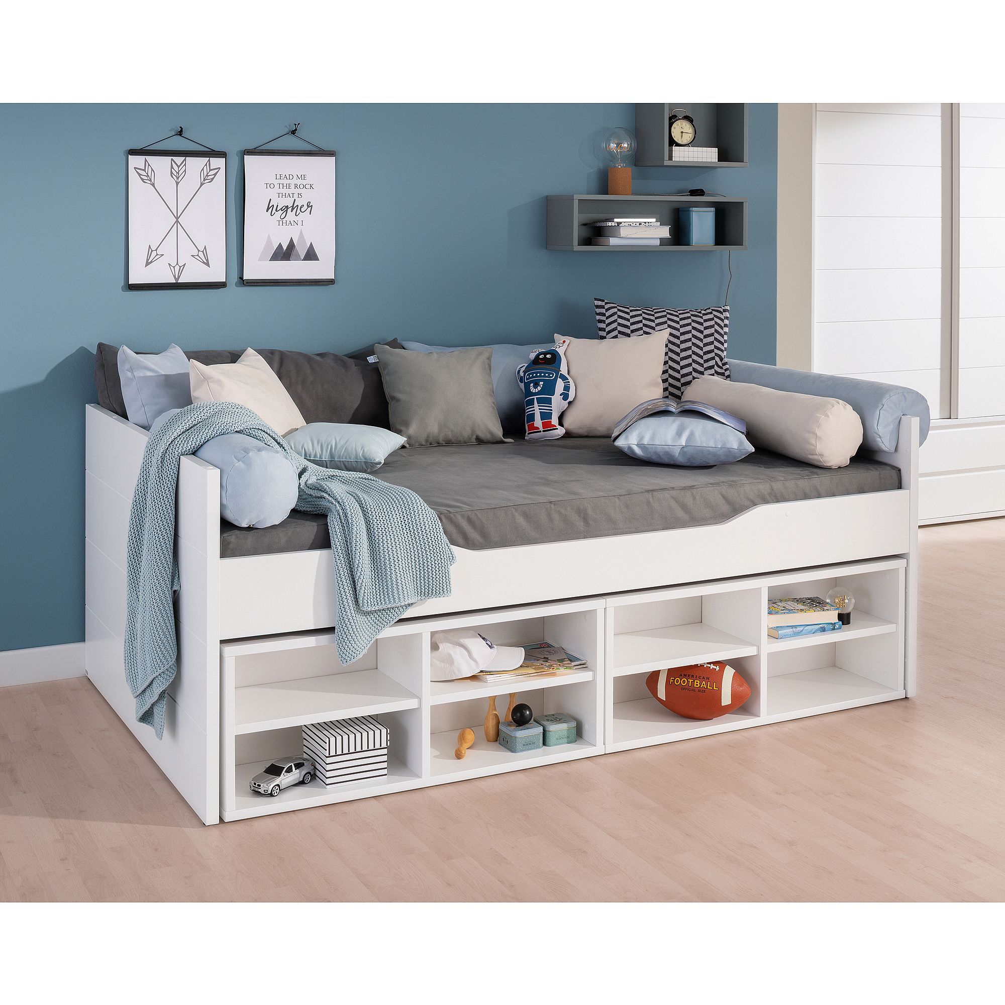 Weißes Tagesbett mit grauer Bettwäsche, Kissen, Regalen und Deko in einem modernen Zimmer.