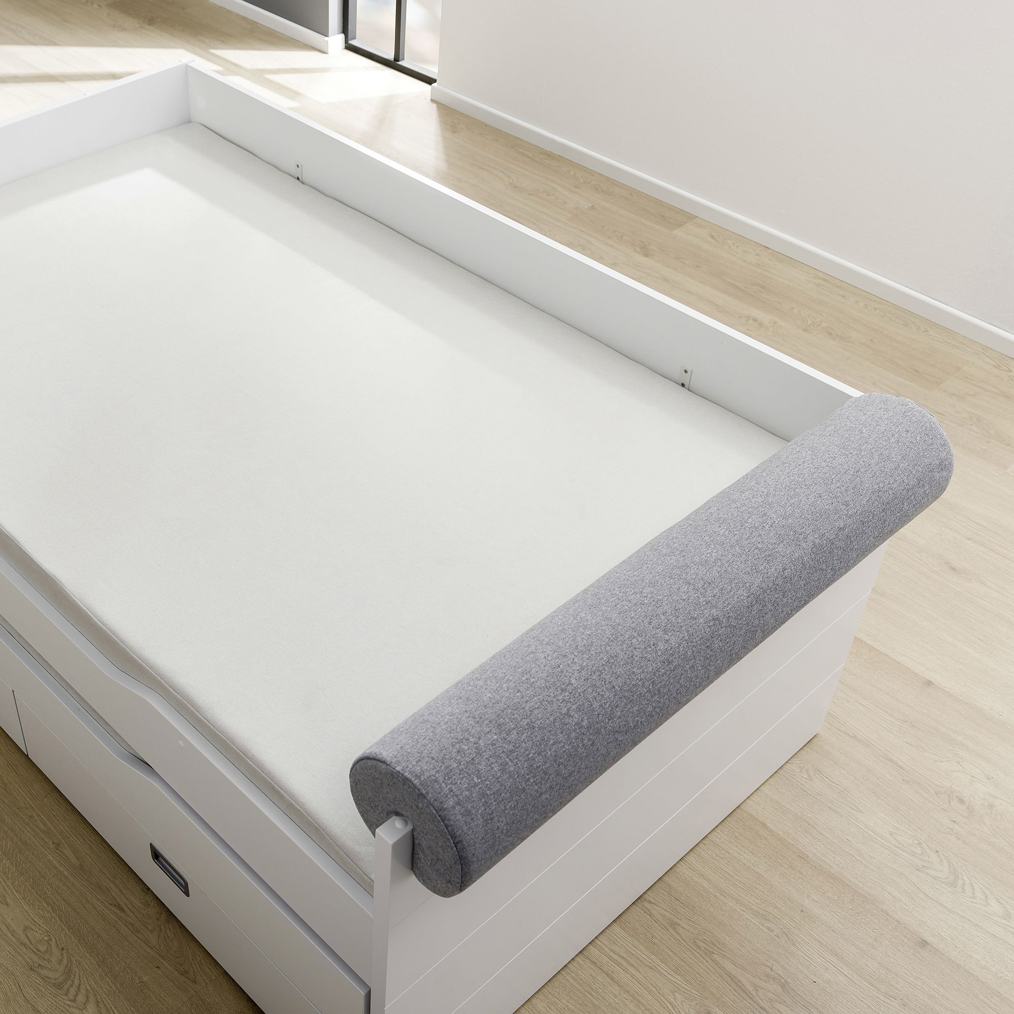 White bed with a gray cylindrical cushion at the head and light wood flooring.