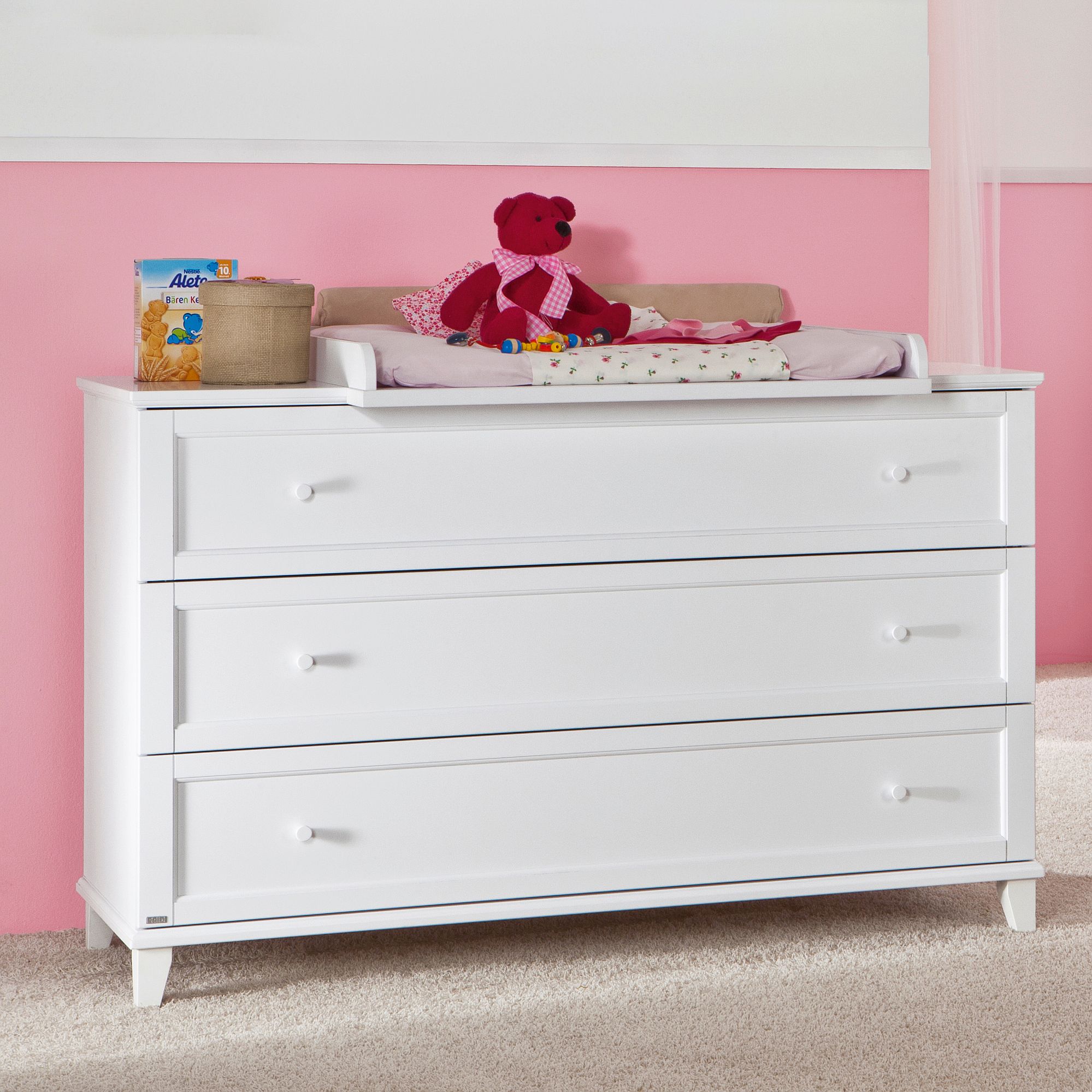 White dresser with changing pad, red teddy bear, and baby items against a pink wall.