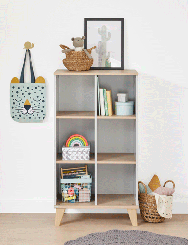 A small bookshelf with books, toys, and baskets, beside a wall with a hanging cat-face bag and woven baskets on the floor.