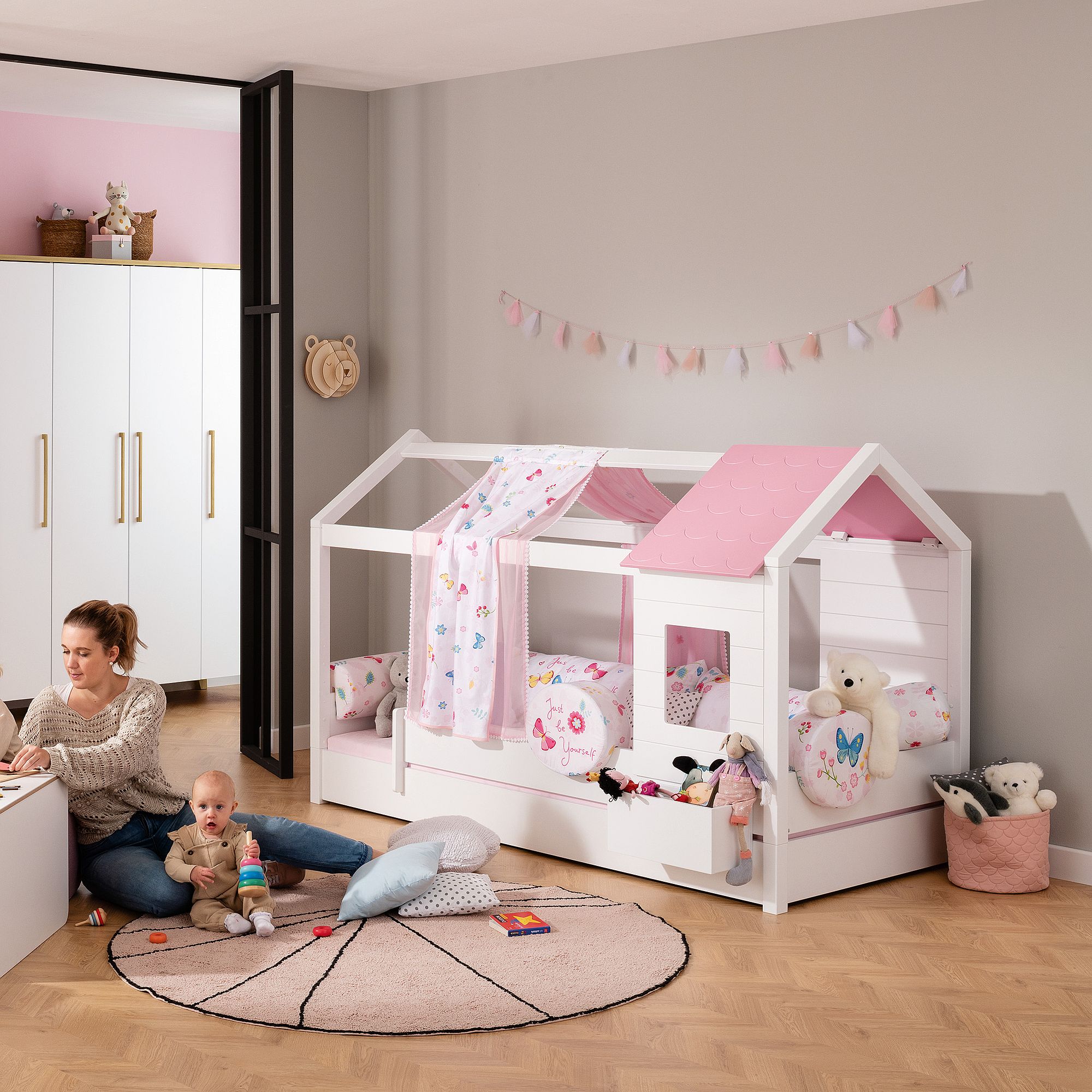 Woman and baby play in a pink and white kids’ room with a house-shaped bed.