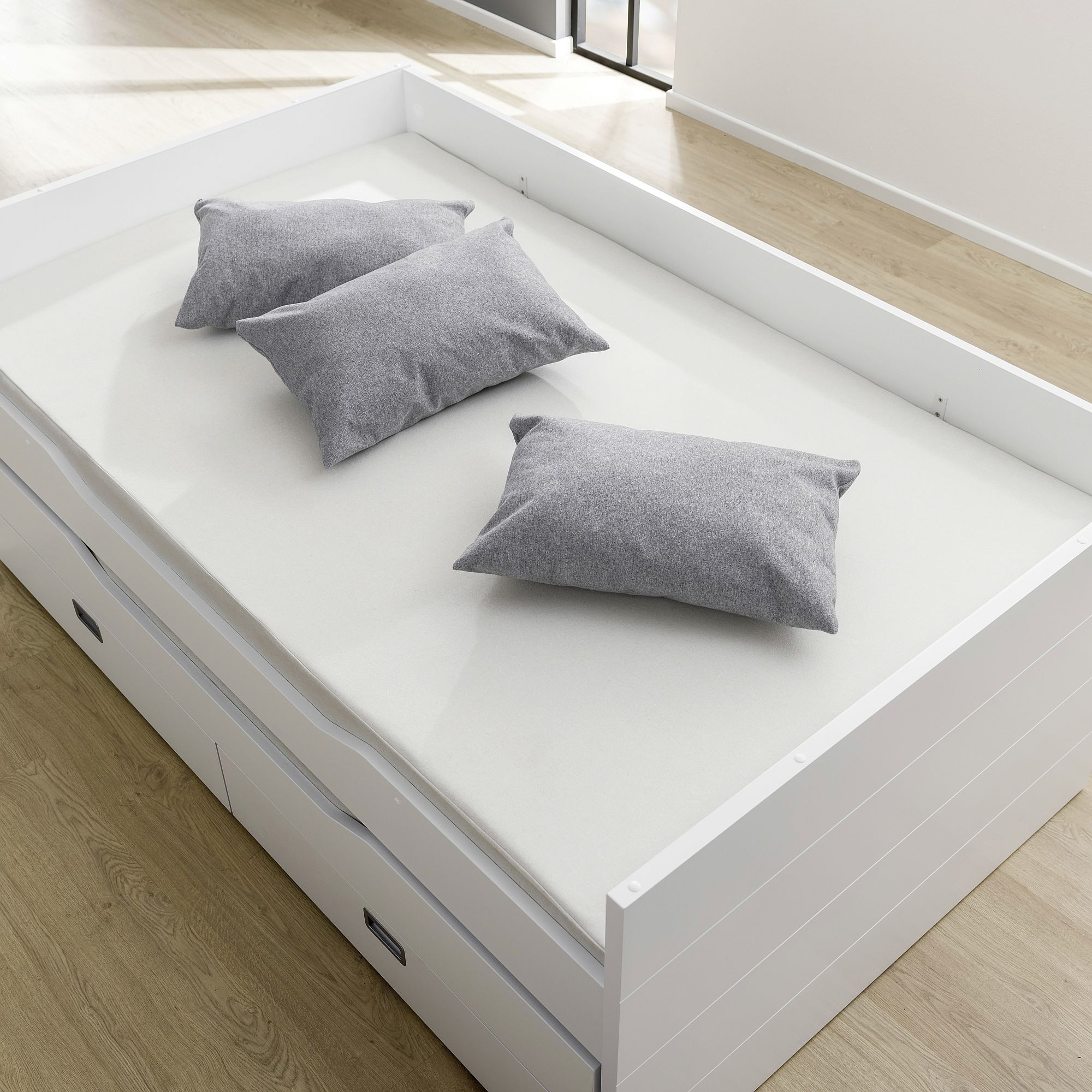 A white bed with three gray pillows on a light wood floor in a bright room.