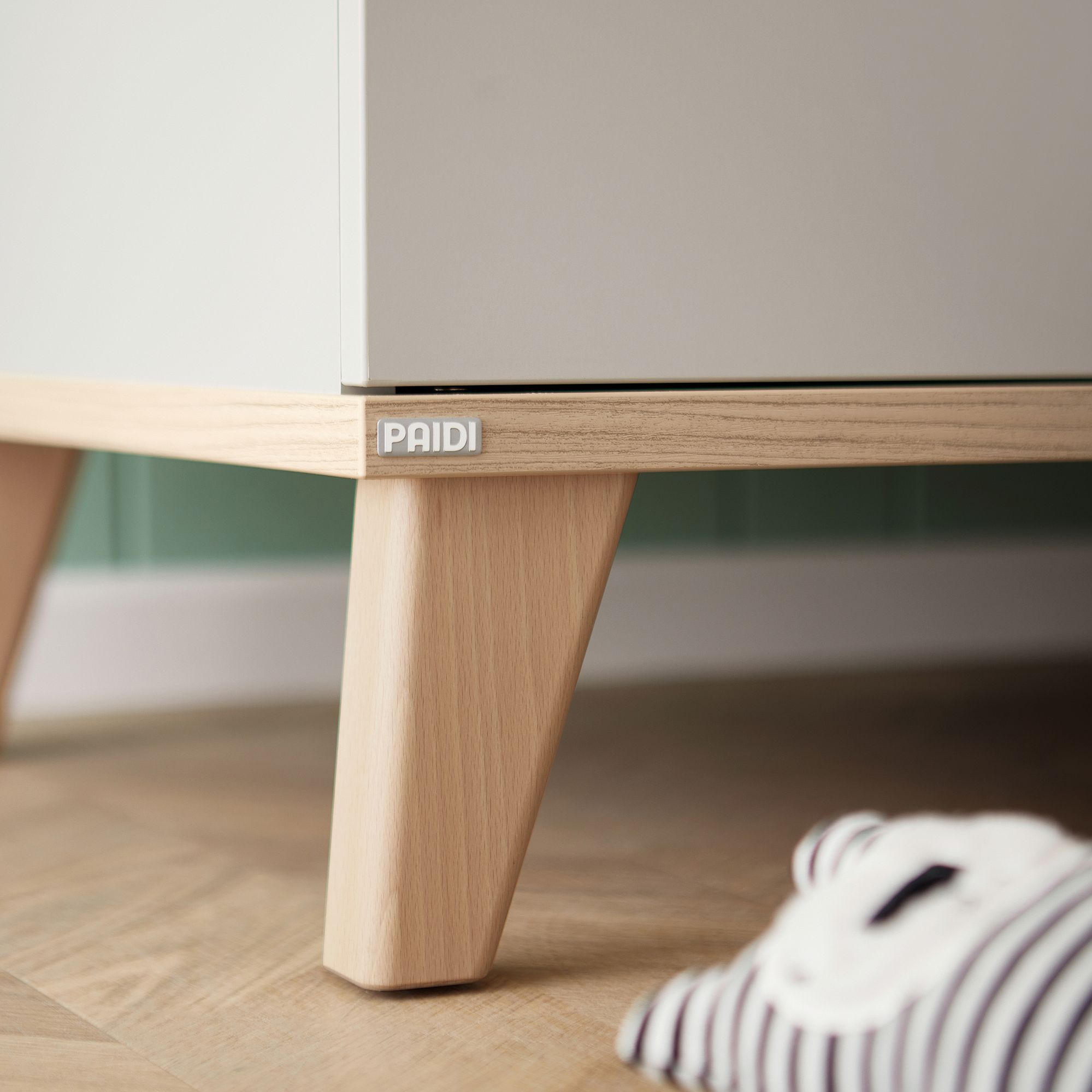 Standregal Fiene mit 2 Türen Nahaufnahme eines Holzmöbelbeins mit PAIDI-Etikett, gestreifter Stoff im Vordergrund.