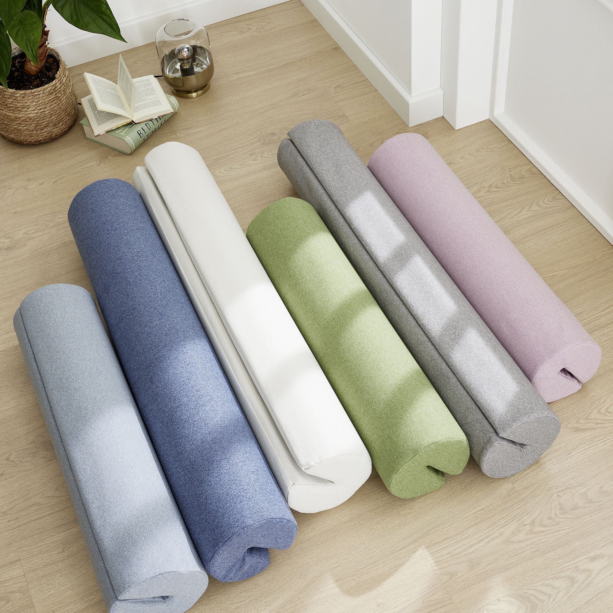 Six rolled yoga mats in pastel colors on a light wood floor near a potted plant.