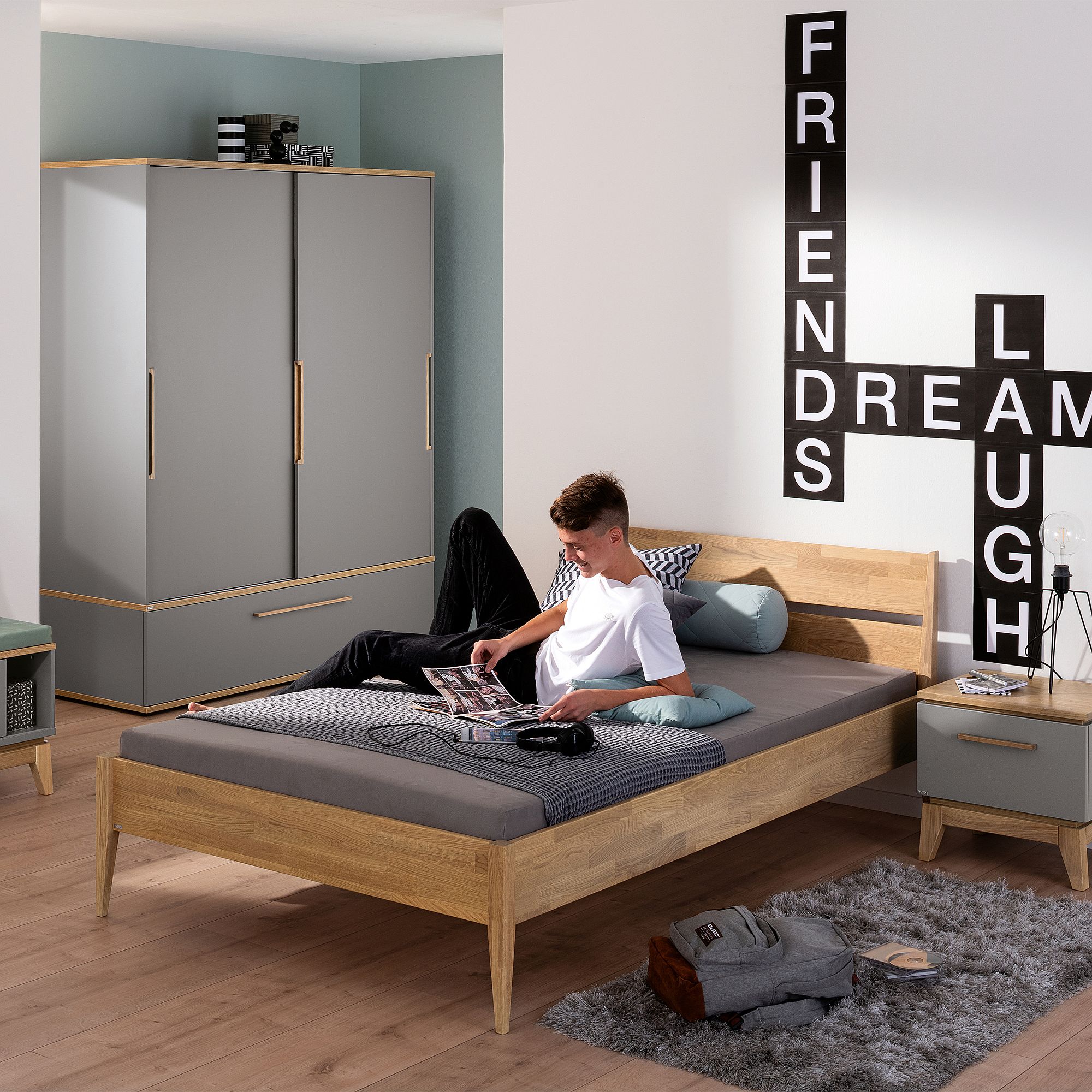 Ein Teenager sitzt mit seinem Laptop auf dem Bett in einem modernen Schlafzimmer, an dessen Wand ein Word-Art-Bild hängt.