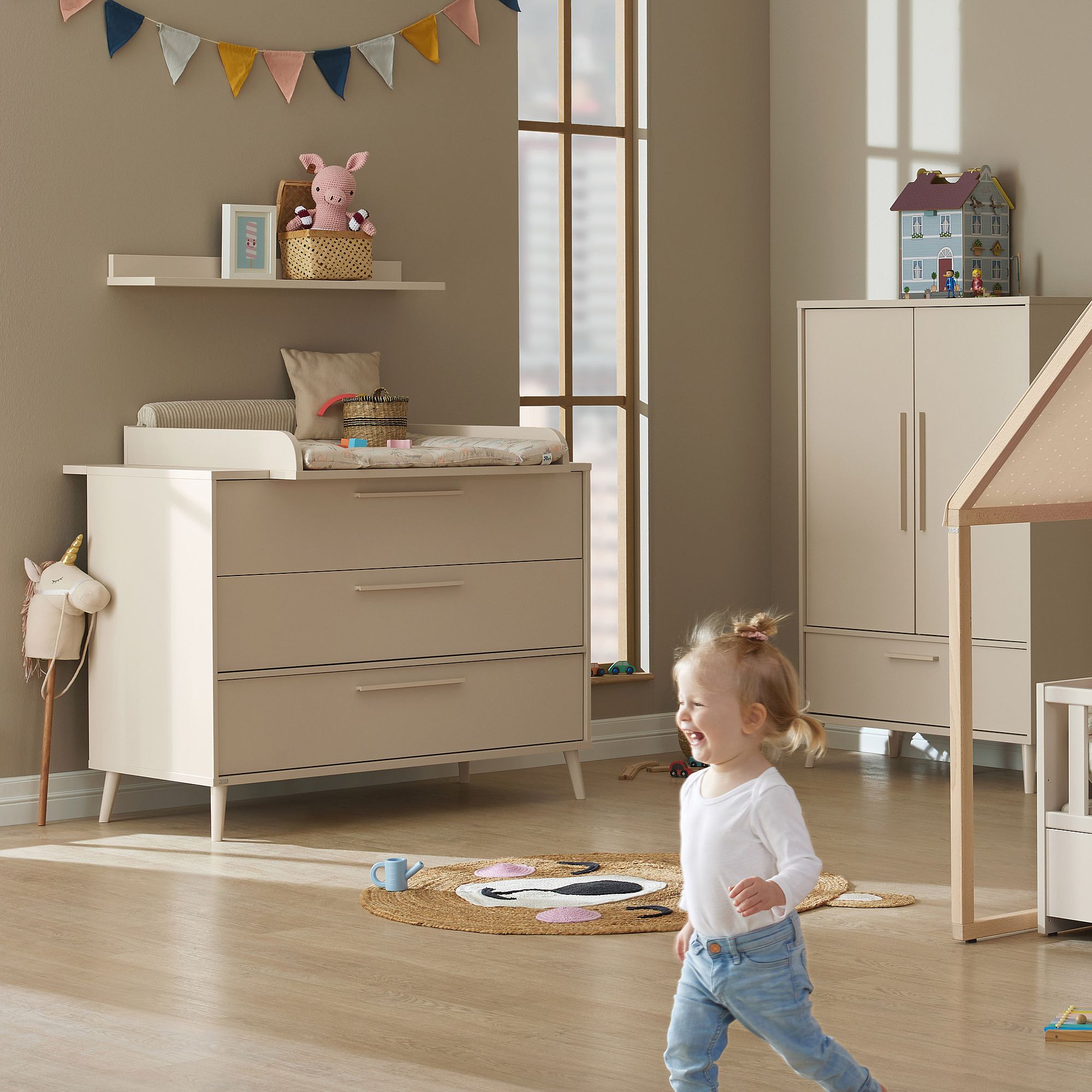 Ein Kleinkind rennt durch einen sonnendurchfluteten Kinderraum mit neutralen Möbeln und verspielter Einrichtung.
