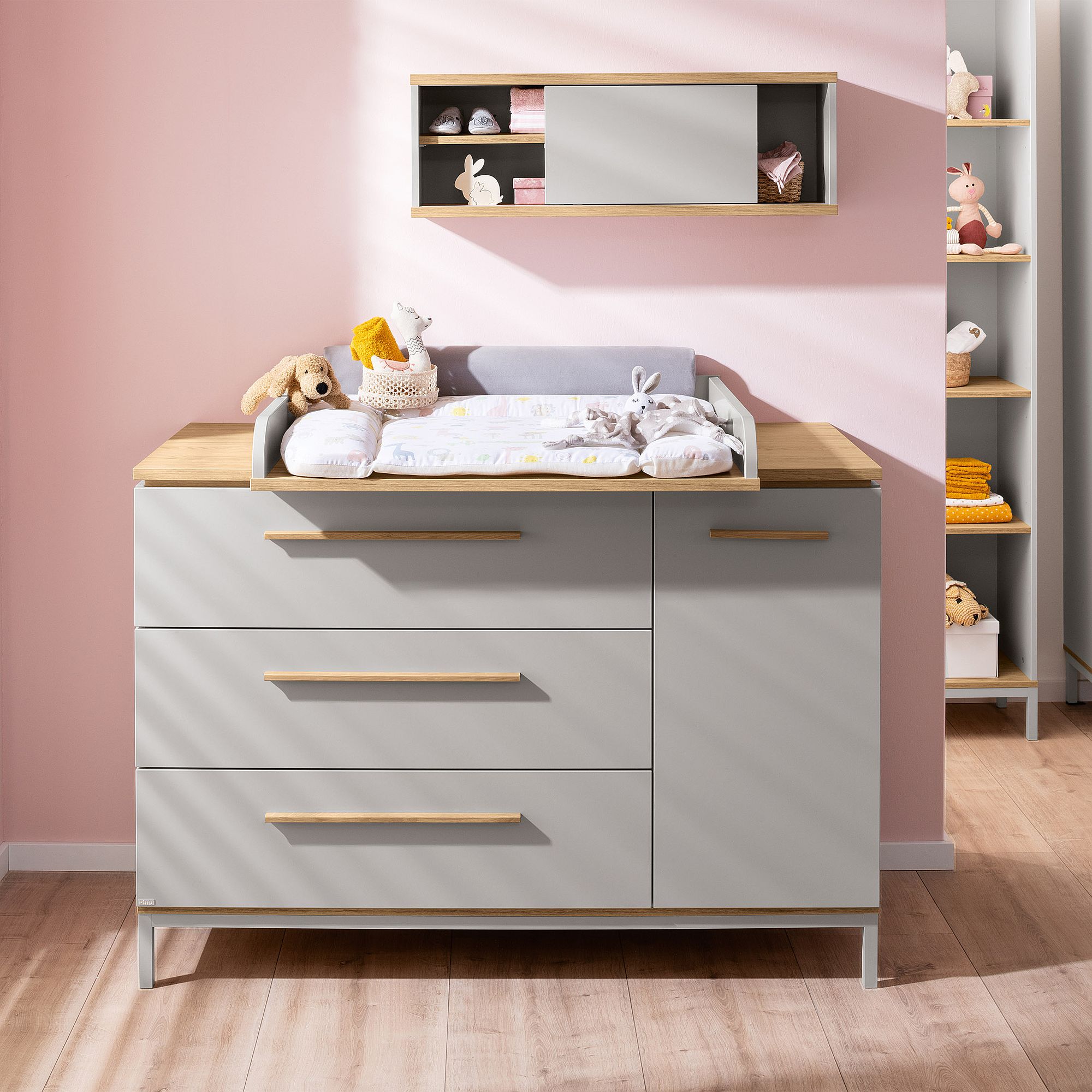 Grauer Wickeltisch mit Kuscheltieren und Babyzubehör vor einer rosa Wand.