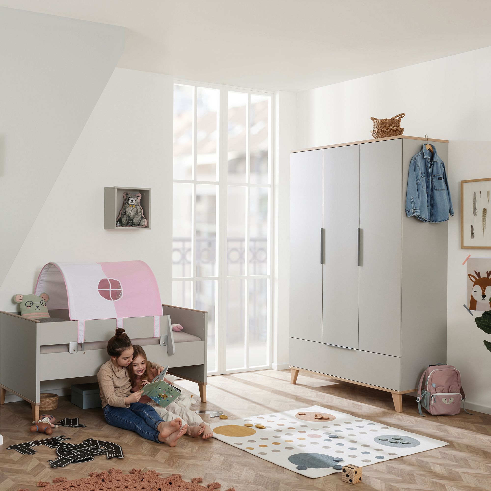 Zwei Kinder lesen auf dem Boden in einem hellen, modernen Kinderzimmer mit Spielzeug und Möbeln.