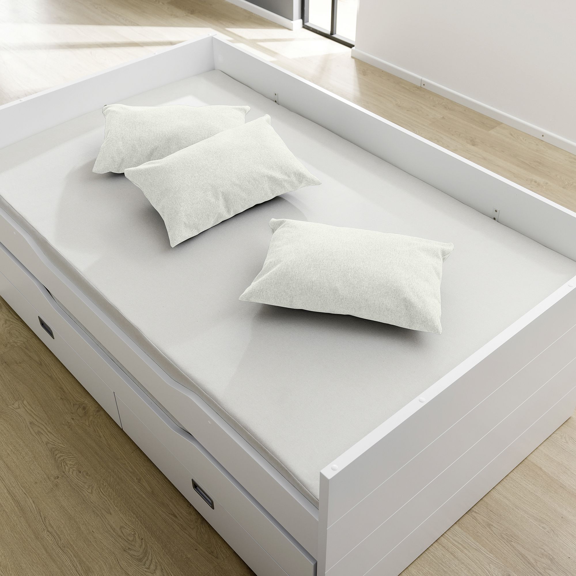 A white bed with three pillows, drawers underneath, and a wooden floor.
