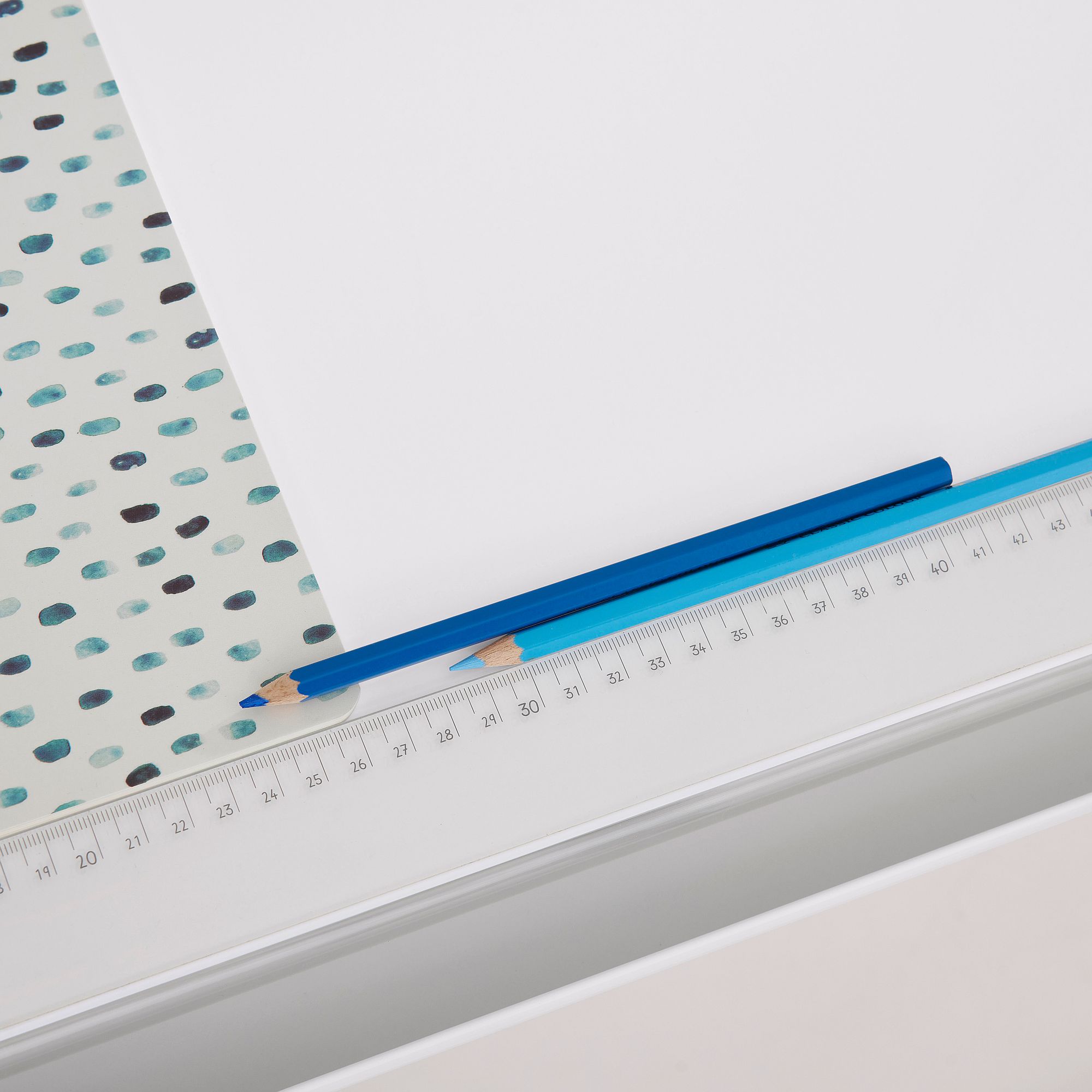 Schülerschreibtisch Diego Curved 120 x 70 cm in Kreideweiß Zwei blaue Bleistifte und ein Lineal auf einer weißen Fläche mit einem gepunkteten Papiermuster.