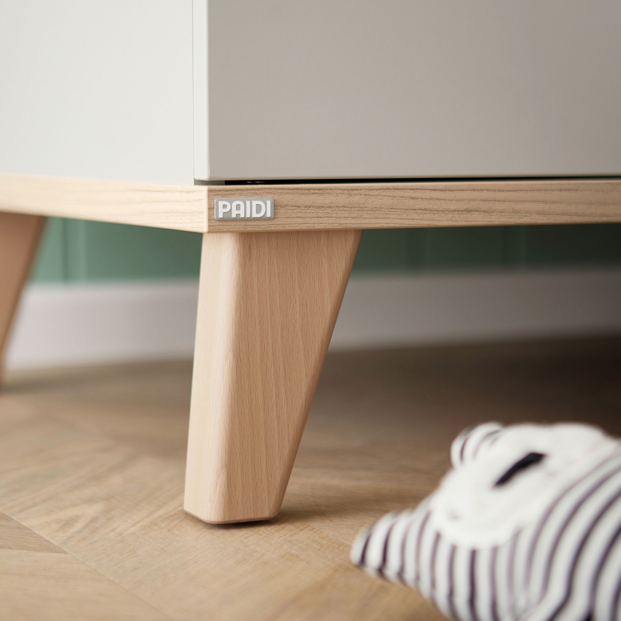 Möbel aus hellem Holz mit PAIDI-Etikett, gestreiftes Plüschtier auf dem Boden daneben.