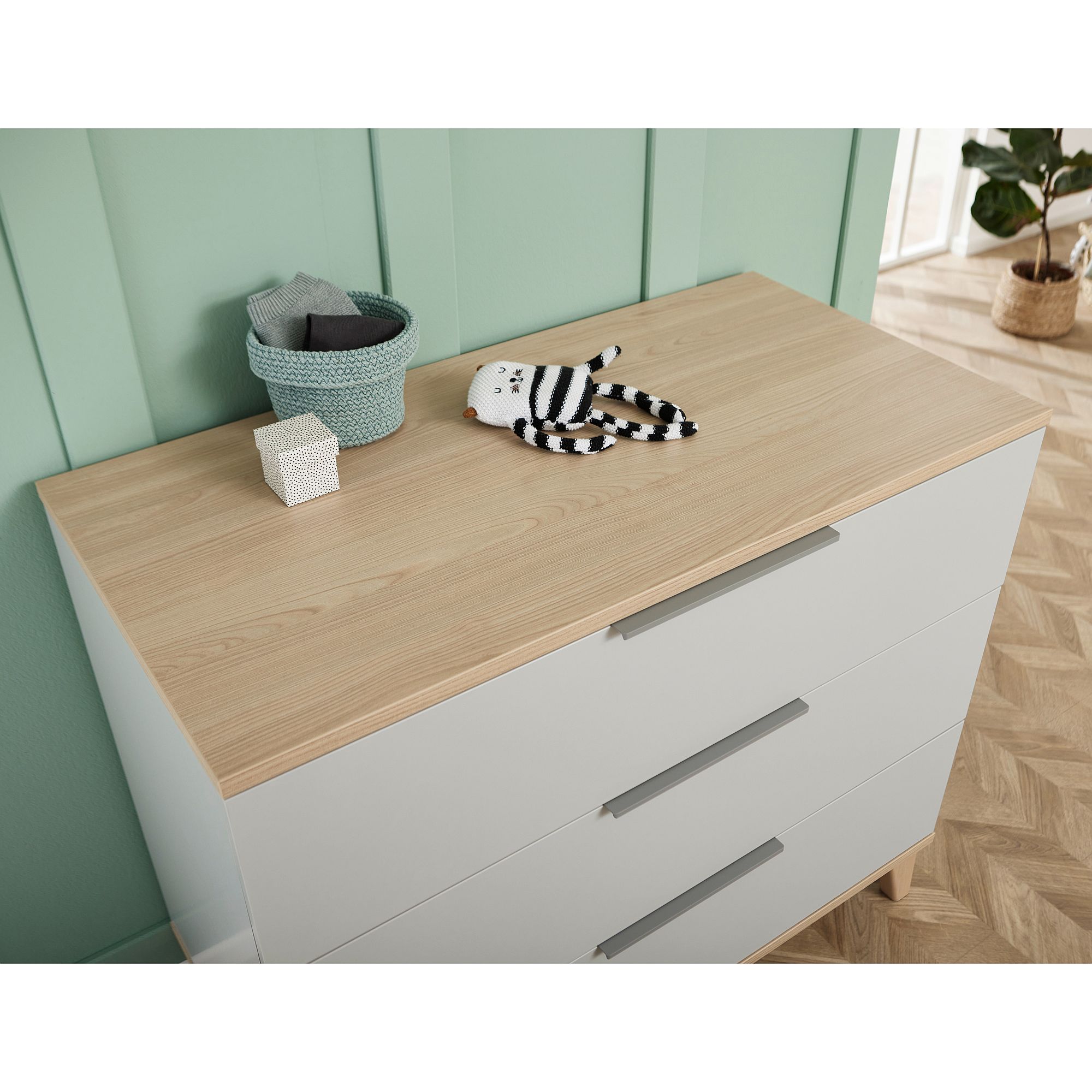 Light wood dresser with basket, toy cat, and small box on top against green wall.