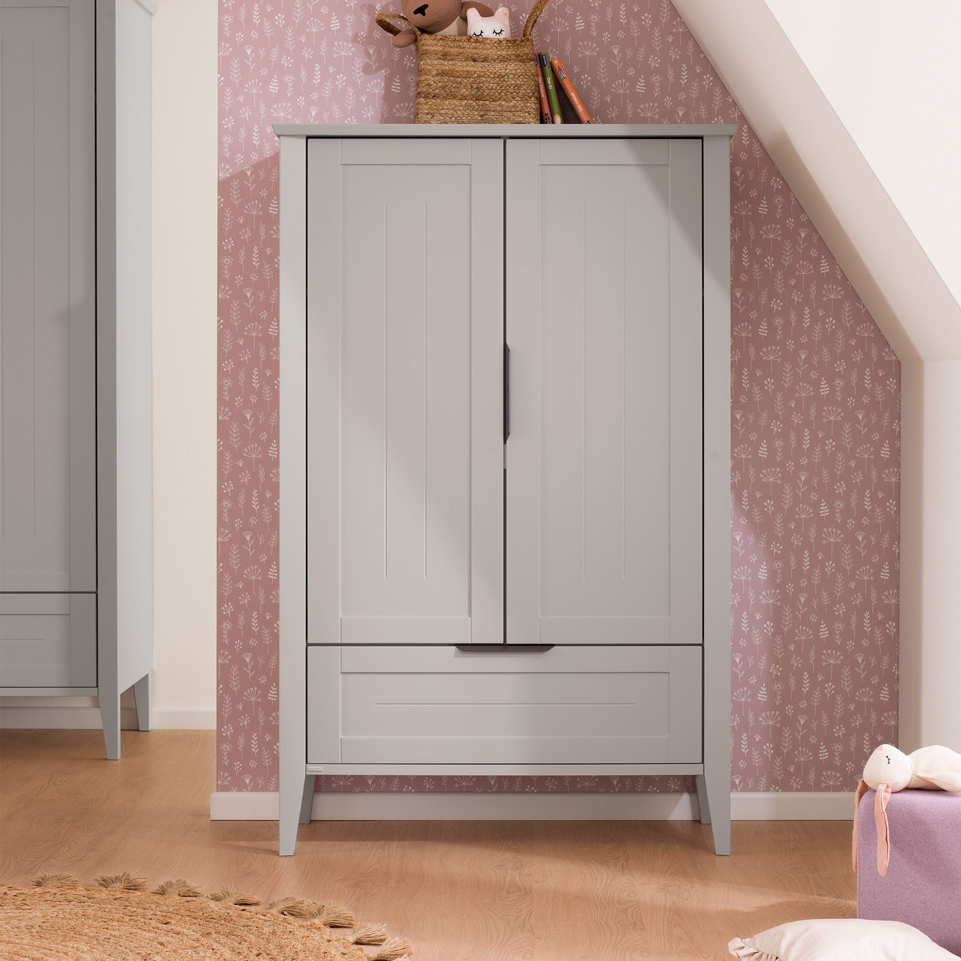 Gray wardrobe with two doors and drawers against pink patterned wallpaper.