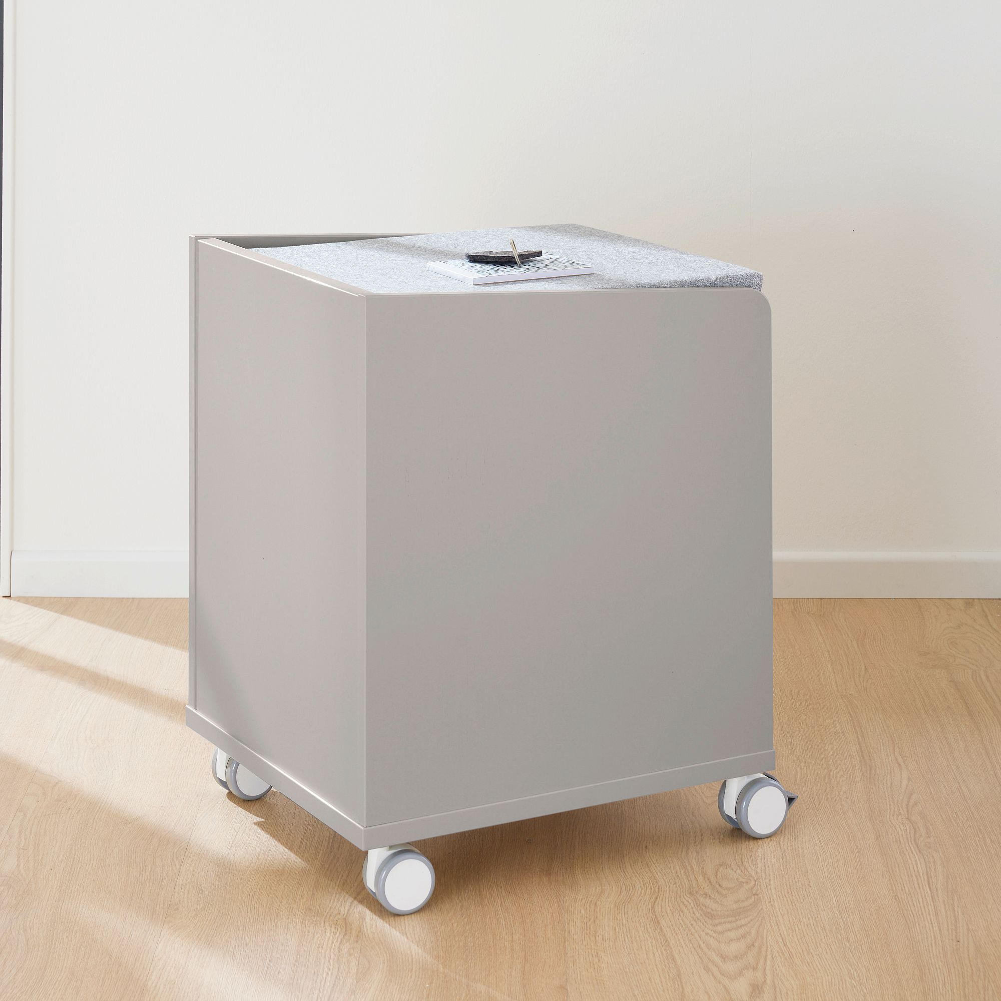 Rollcontainer  mit 2 Schubladen in Kieselgrau Ein grauer Kasten auf Rädern mit einem Filzdeckel, der auf einem Holzboden steht.