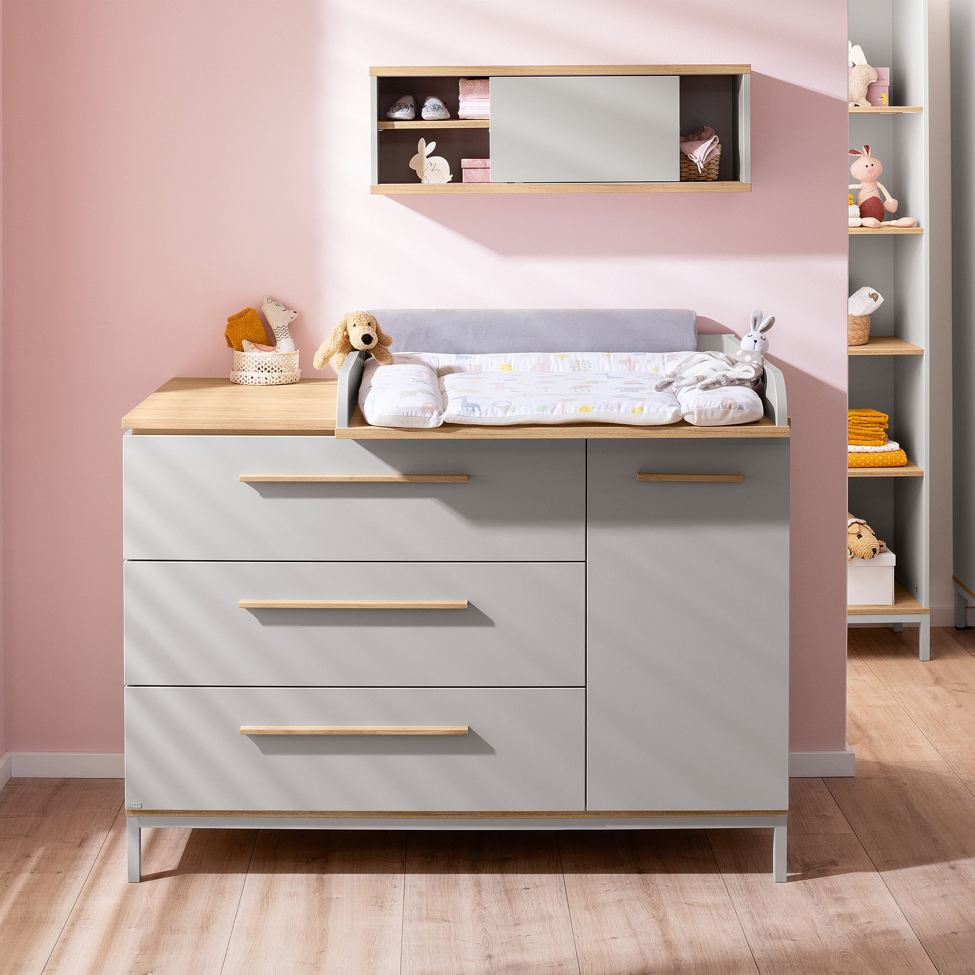 Eine moderne Kinderzimmer-Kommode mit Wickeltisch, Spielzeug und Regalen vor einer rosa Wand.