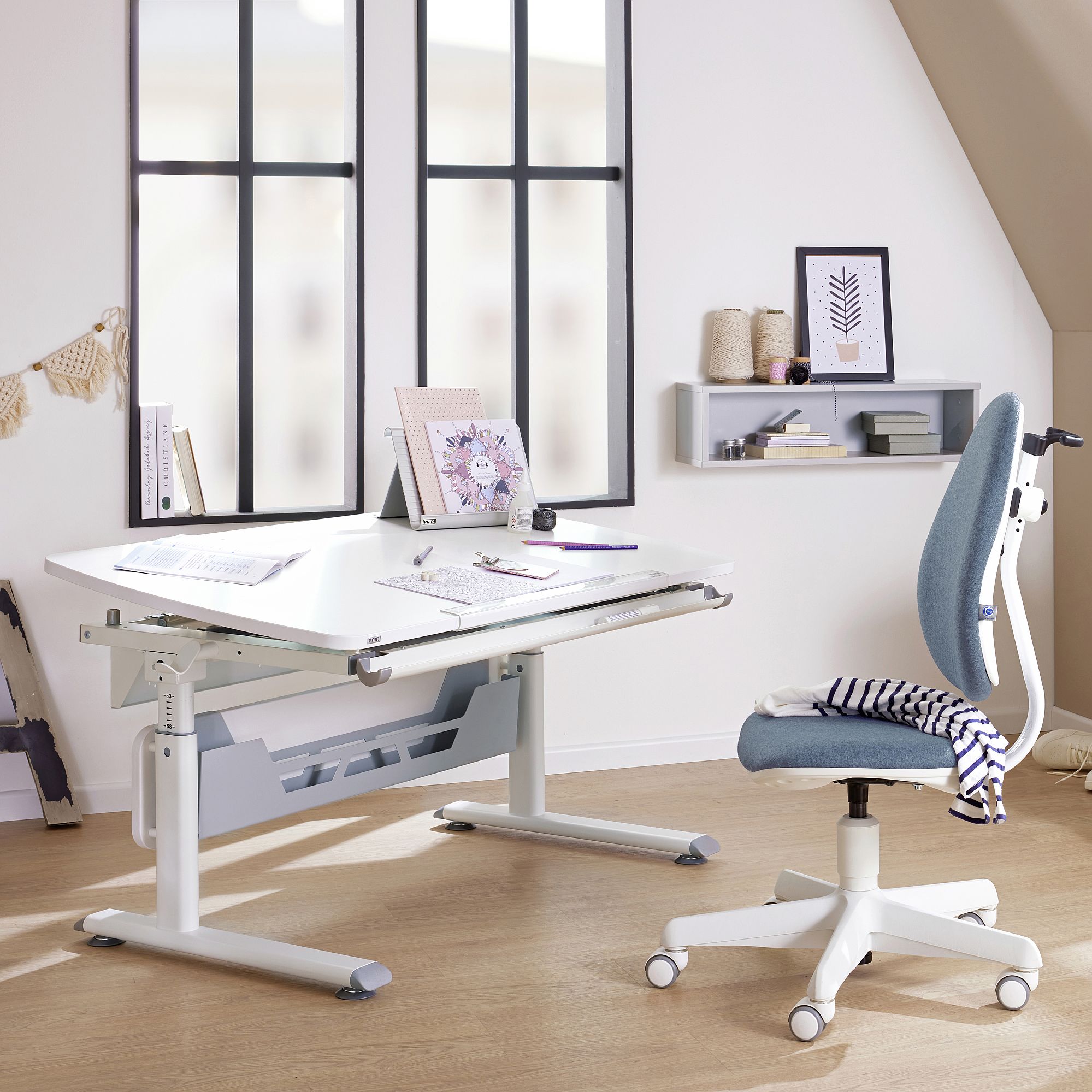 Schülerschreibtisch Diego Curved 120 x 70 cm in Kreideweiß Moderner Arbeitsbereich mit kippbarem Schreibtisch, blauem Stuhl und Wandregalen an den Fenstern.