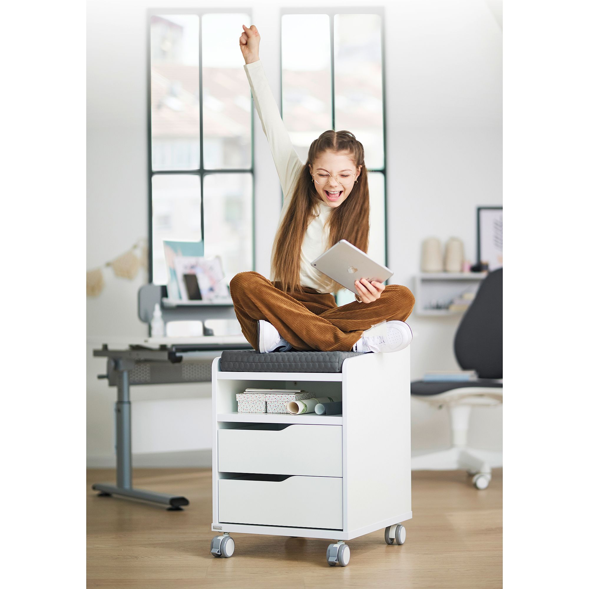 Girl sitting on rolling cabinet, cheering, holding a tablet in a bright room.