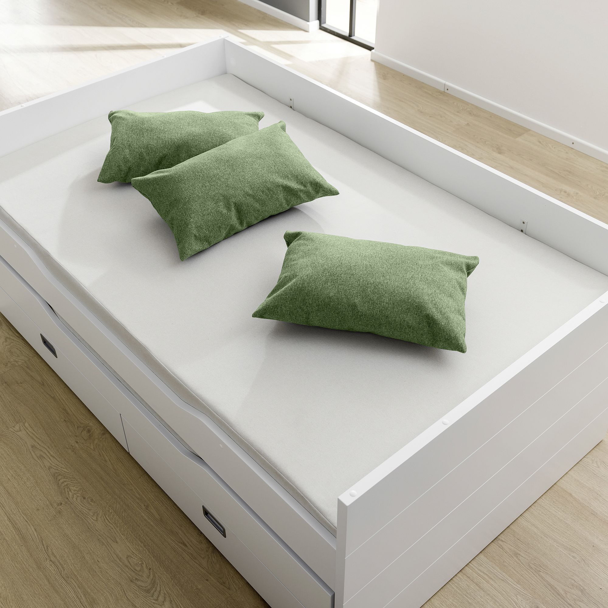 White bed with three green pillows on a light wood floor in a bright room.