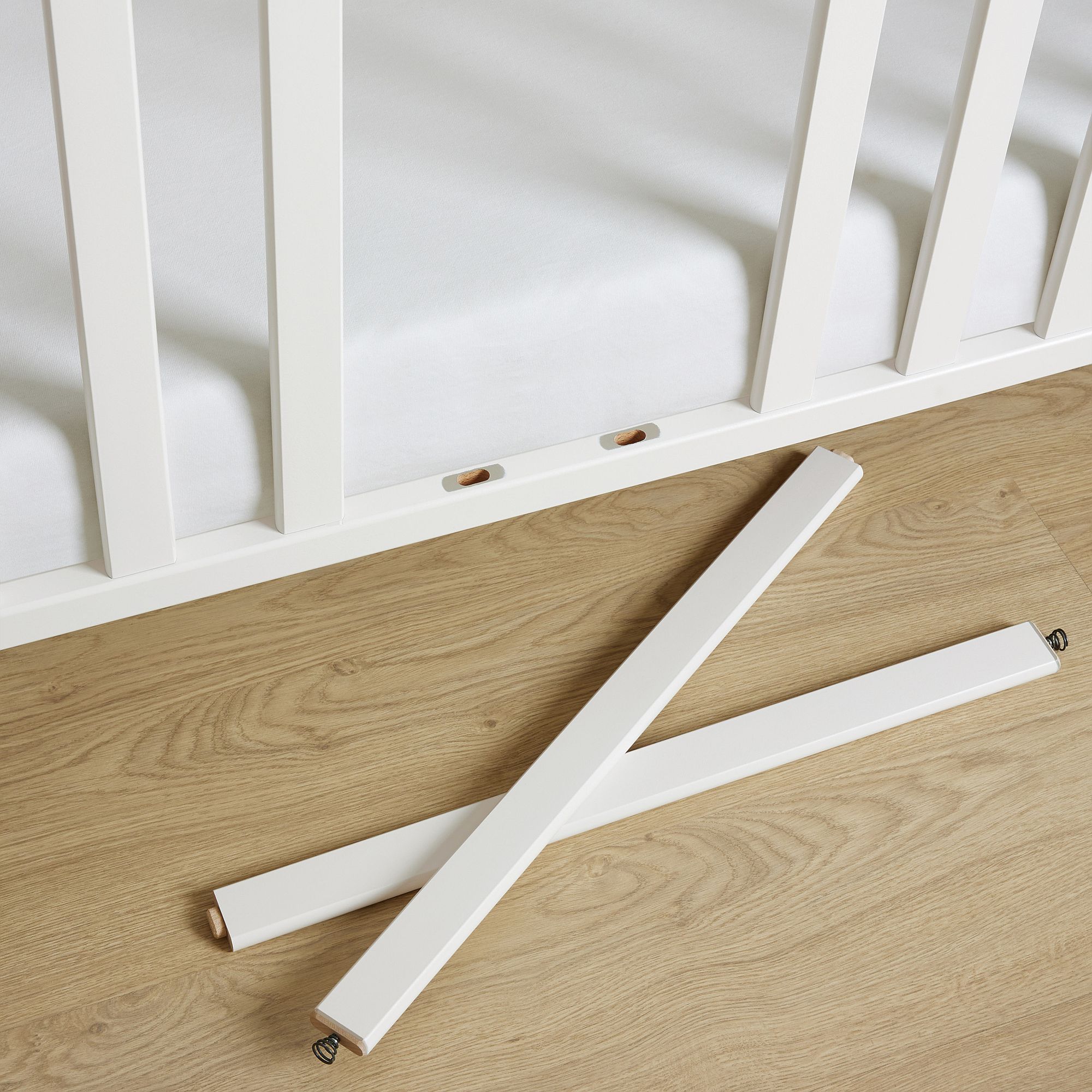 Weißes Kinderbett mit zwei abgetrennten weißen Stäben, die auf einem Holzboden liegen.