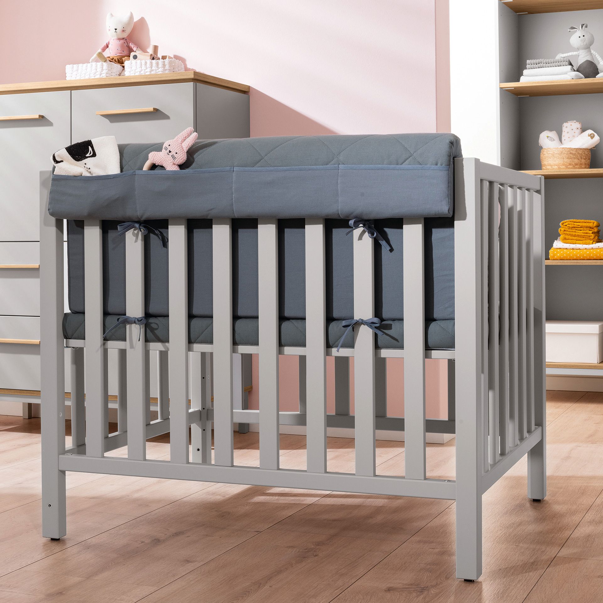 A gray baby crib with a blue bumper in a softly lit nursery room.