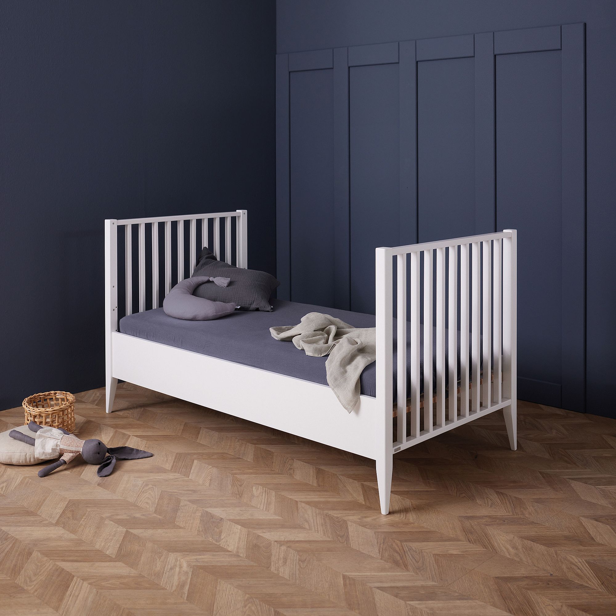 Weißes Kleinkindbett mit grauer Bettwäsche in einem Zimmer mit dunkelblauen Wänden und Holzboden.