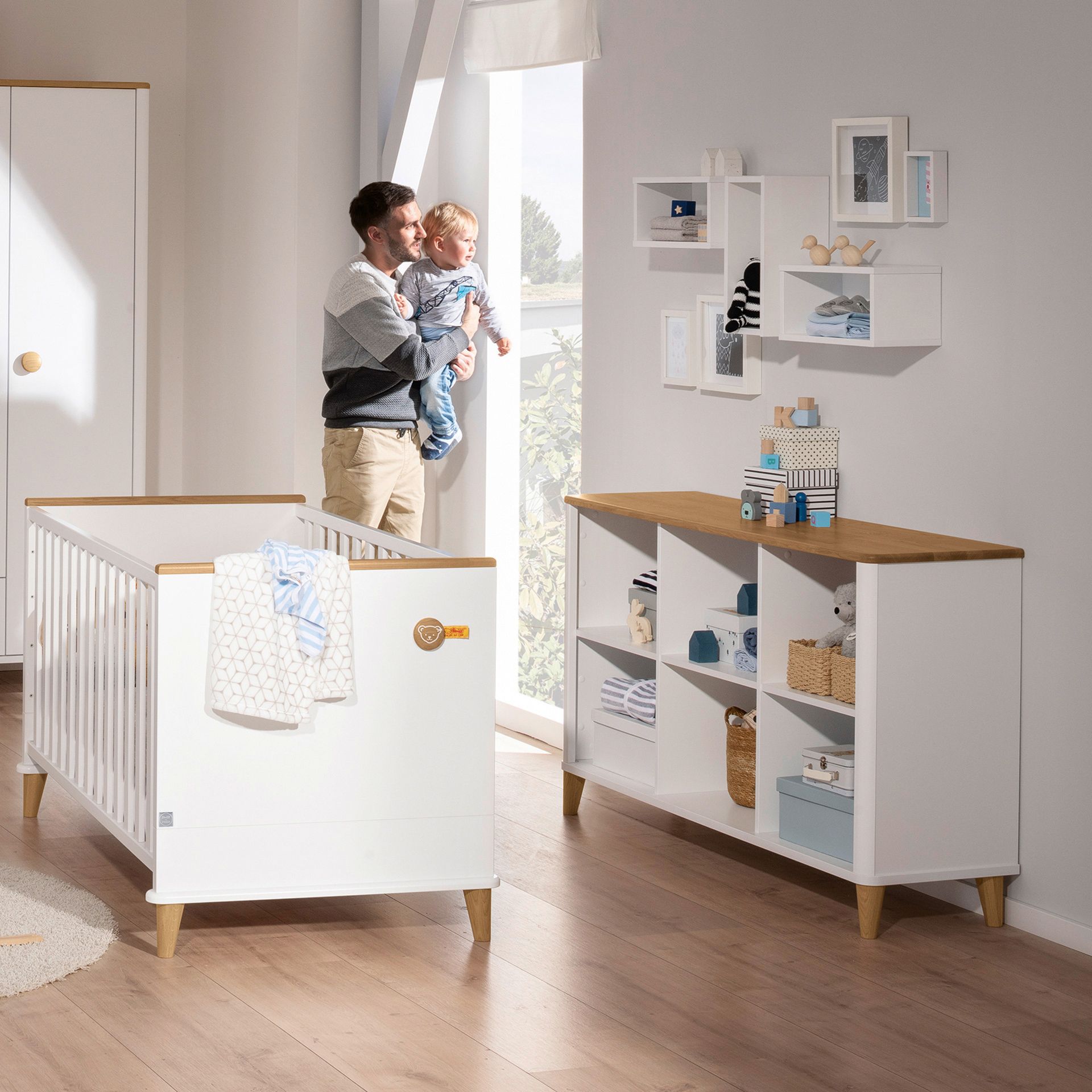 A man holds a baby in a bright, modern nursery with white furniture.