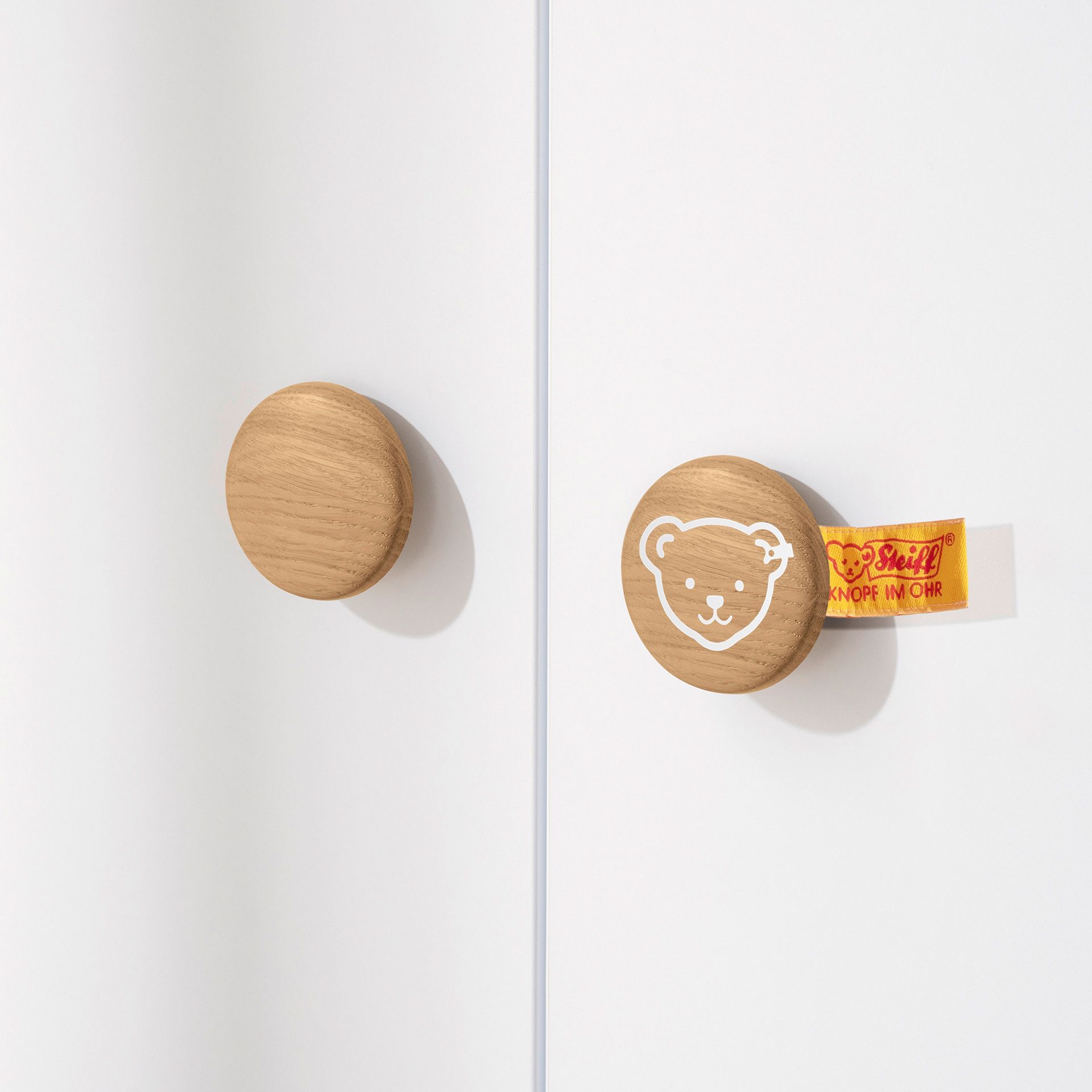 Drehtürenschrank Lotte & Fynn - Steiff by PAIDI mit 3 Türen und 1 Schublade Zwei hölzerne Schrankknöpfe, einer mit einem Bärengesicht und einem gelben Schild dahinter.