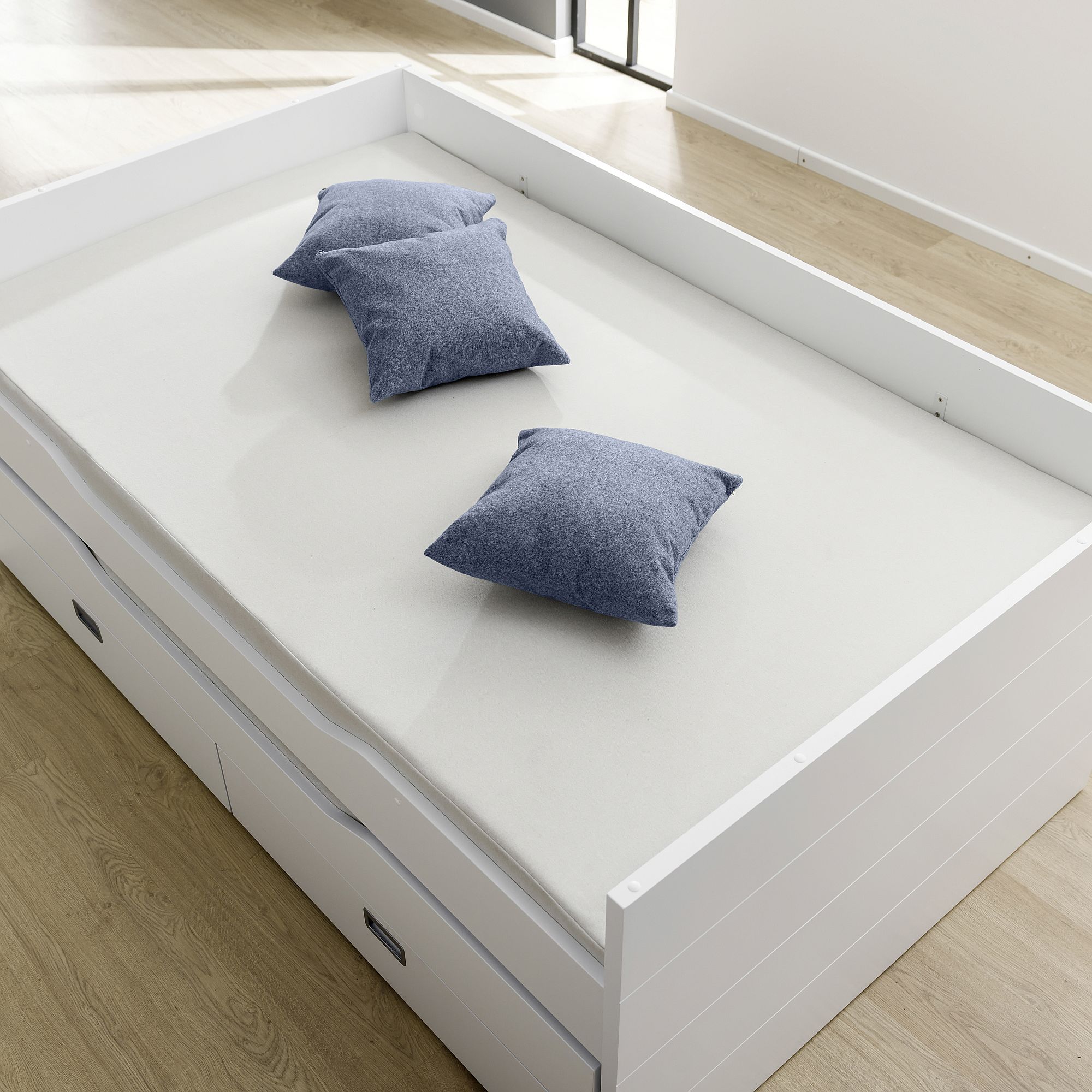 White bed with light bedding and three blue pillows on a wooden floor.