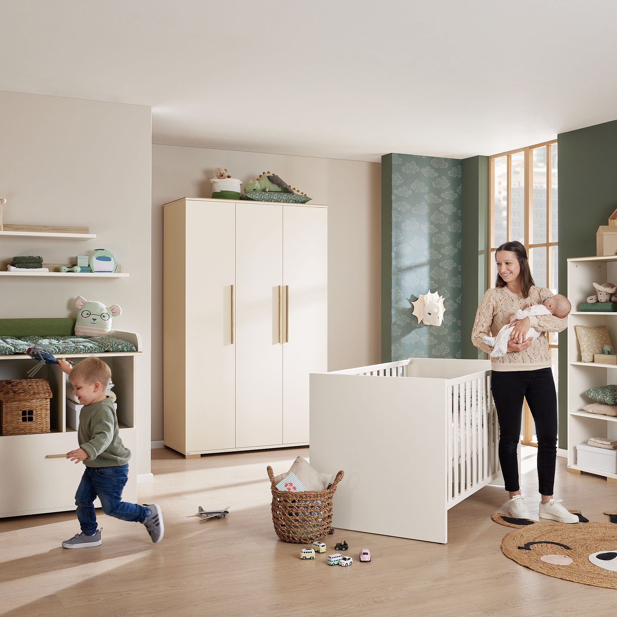 Eine Frau hält ein Baby in einem modernen Kinderzimmer, während ein Kleinkind in der Nähe rennt.