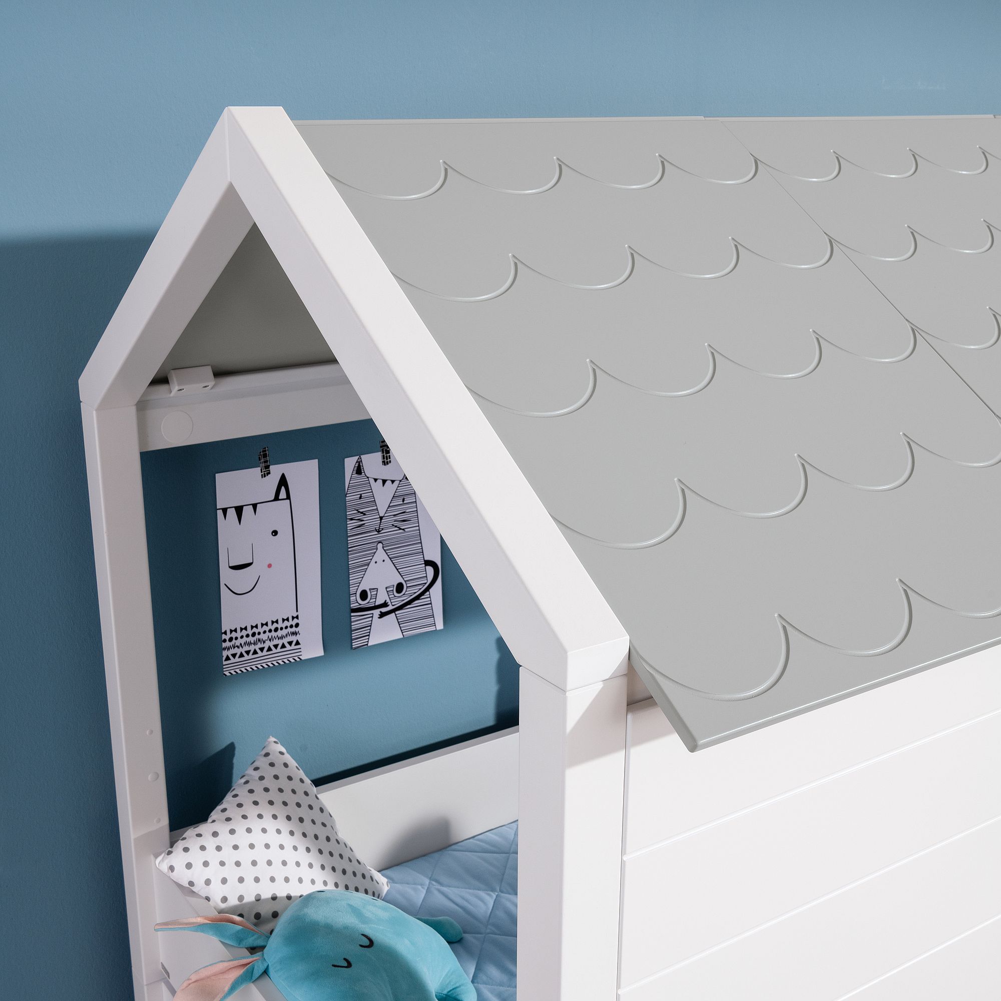 Weißes Bett in Hausform mit grauem Dach und verspielter Wandmalerei über blauem Bettzeug.