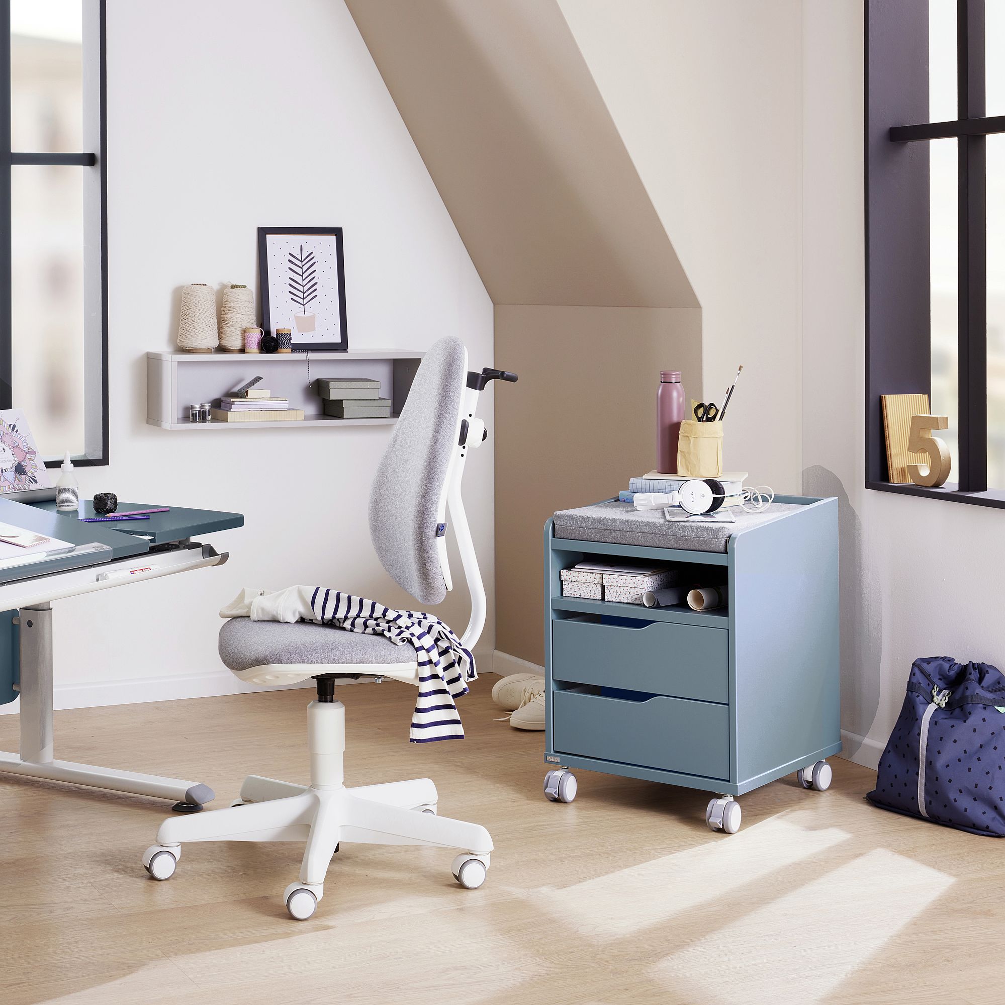 Modernes Arbeitszimmer mit Schreibtisch, Rollstuhl, blauem Schubladenschrank und großen Fenstern.