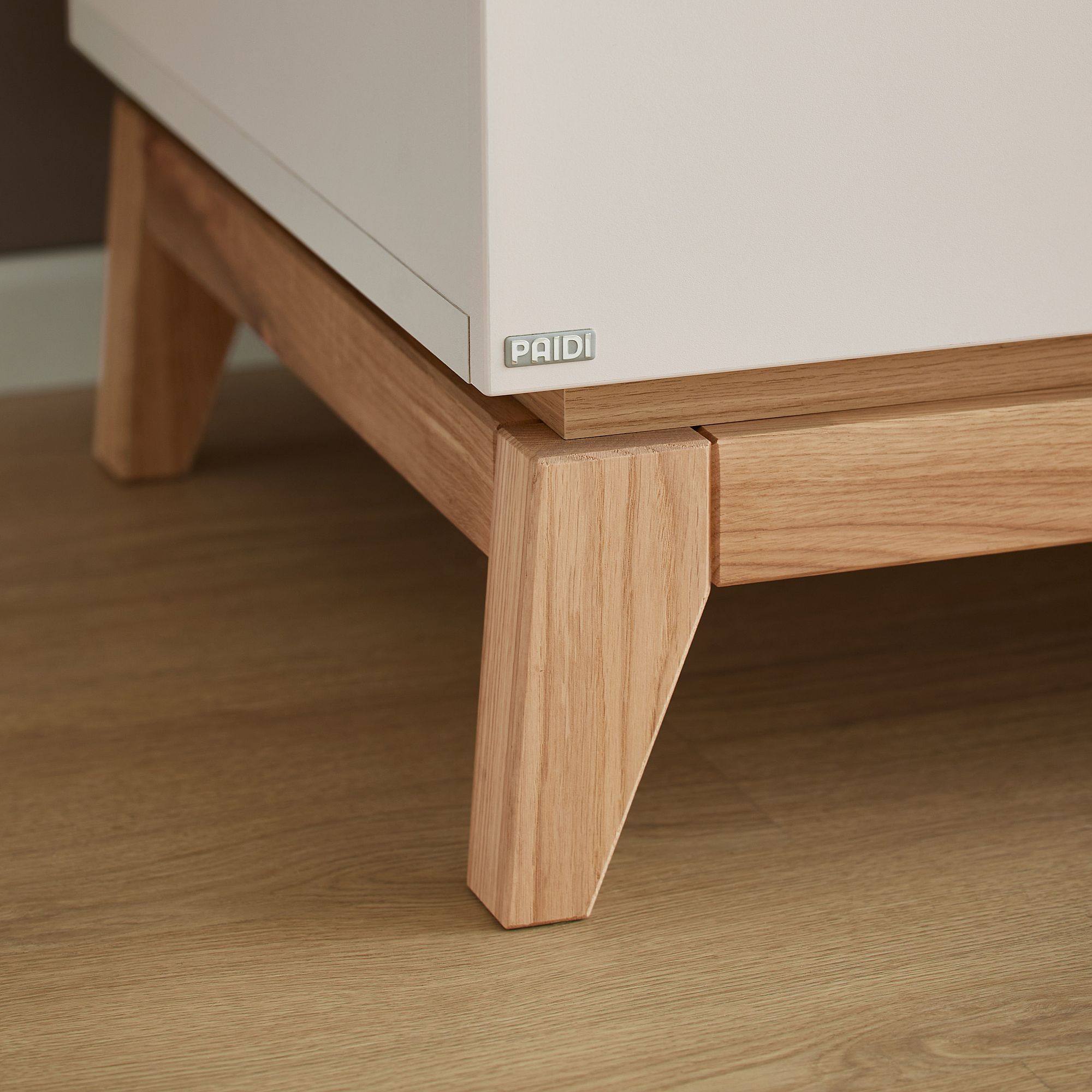 Holzbeine eines weißen Schranks auf einem Holzboden, auf dem ein PAIDI-Etikett zu sehen ist.